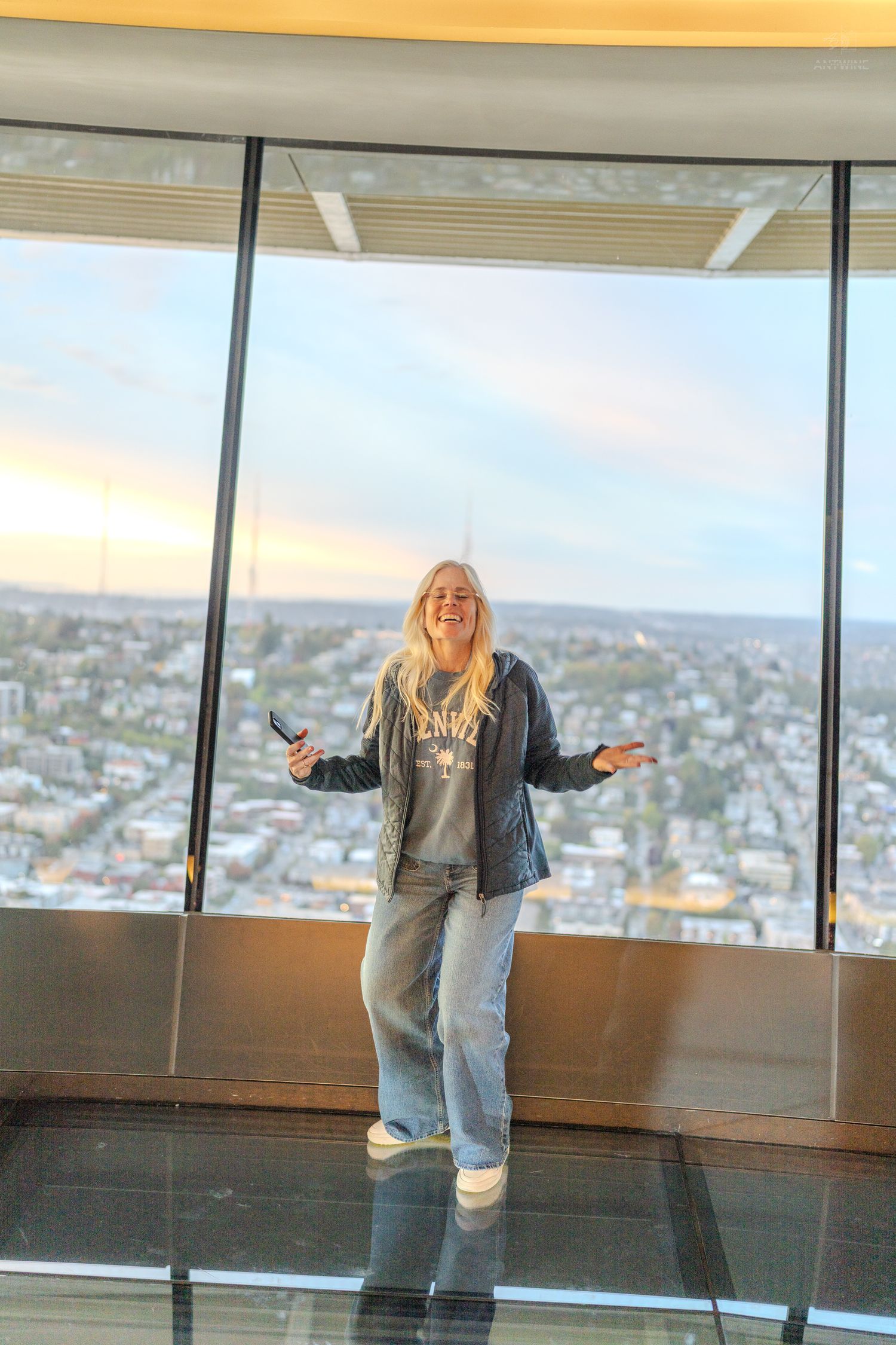 A smiling blonde woman stands arms outstretched on a glass floor observation deck with a panoramic city view at sunset.
