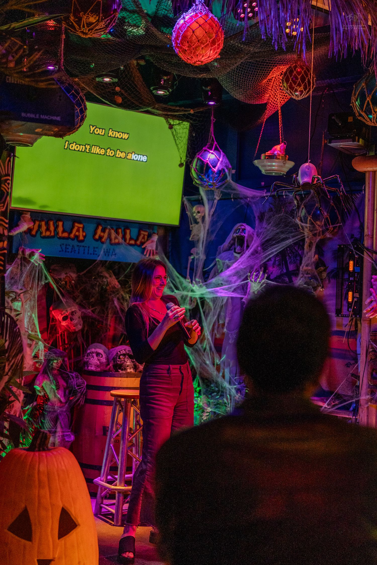 Woman singing karaoke at a Halloween-decorated bar with spiderwebs, skeletons, and a glowing green lyrics screen.