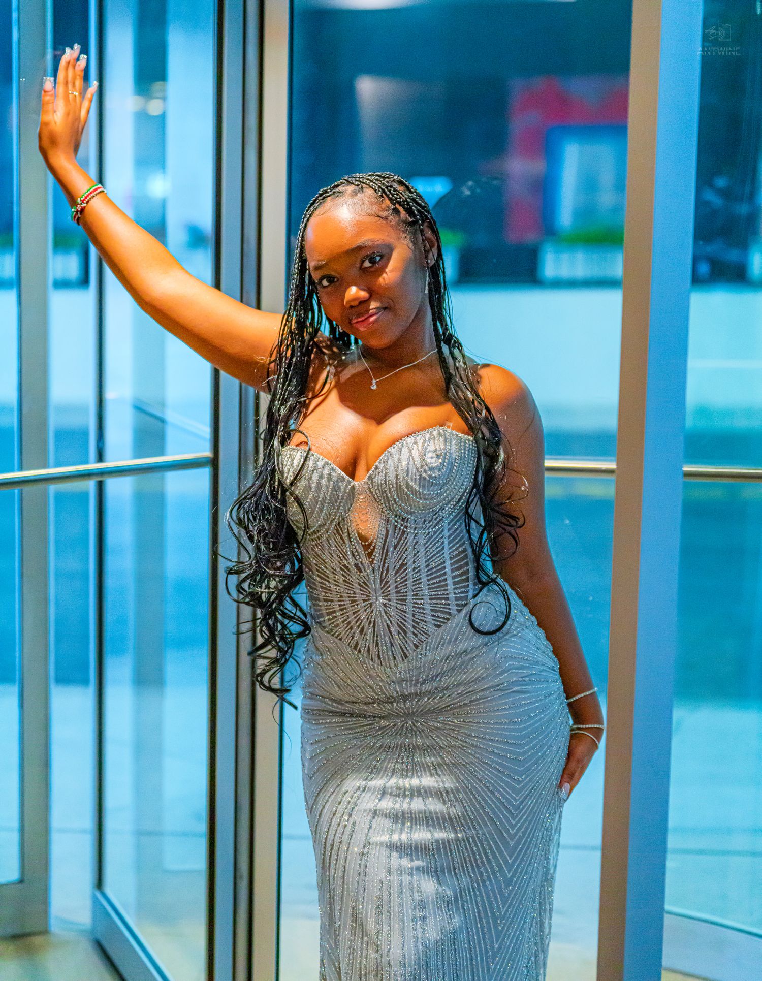 Woman in silver beaded mermaid gown posing elegantly by glass doors, wearing braided hair and waving.