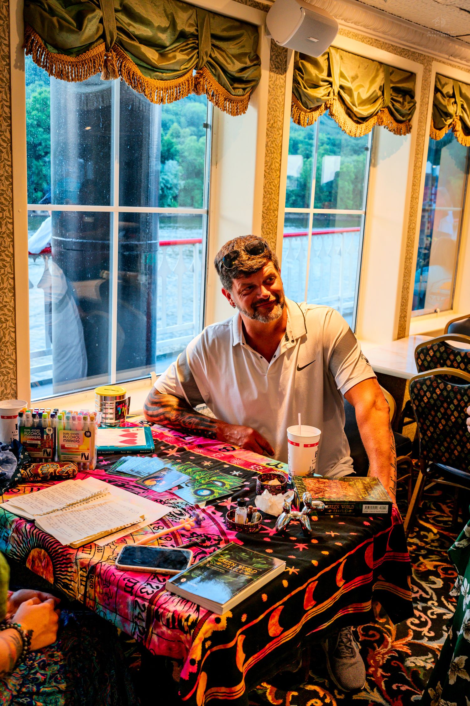 A man in a white Nike polo sits at a colorful table covered with artwork and supplies near bright windows.