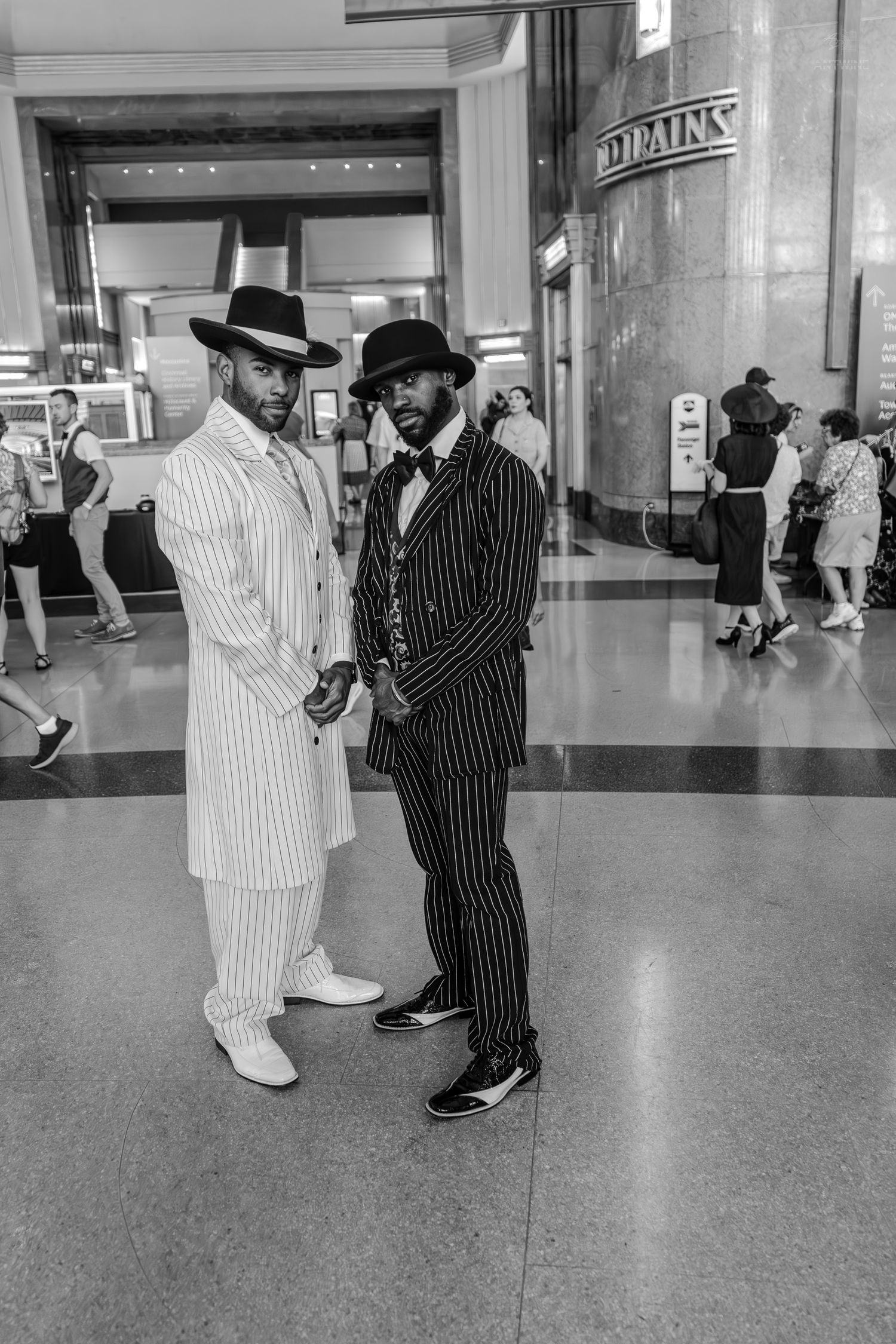 Two stylish men in pinstripe zoot suits and fedora hats pose inside a grand building in black and white photo.