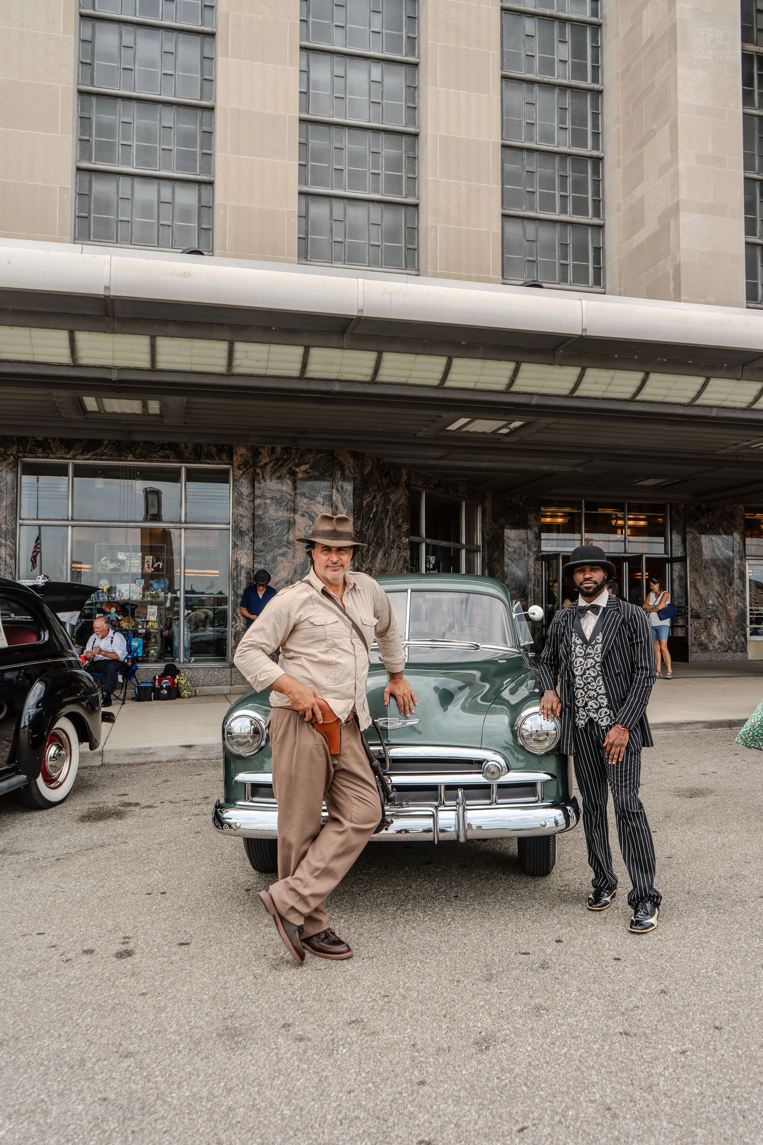 Two men in vintage 1940s costumes pose beside a classic green car outside a historic building.