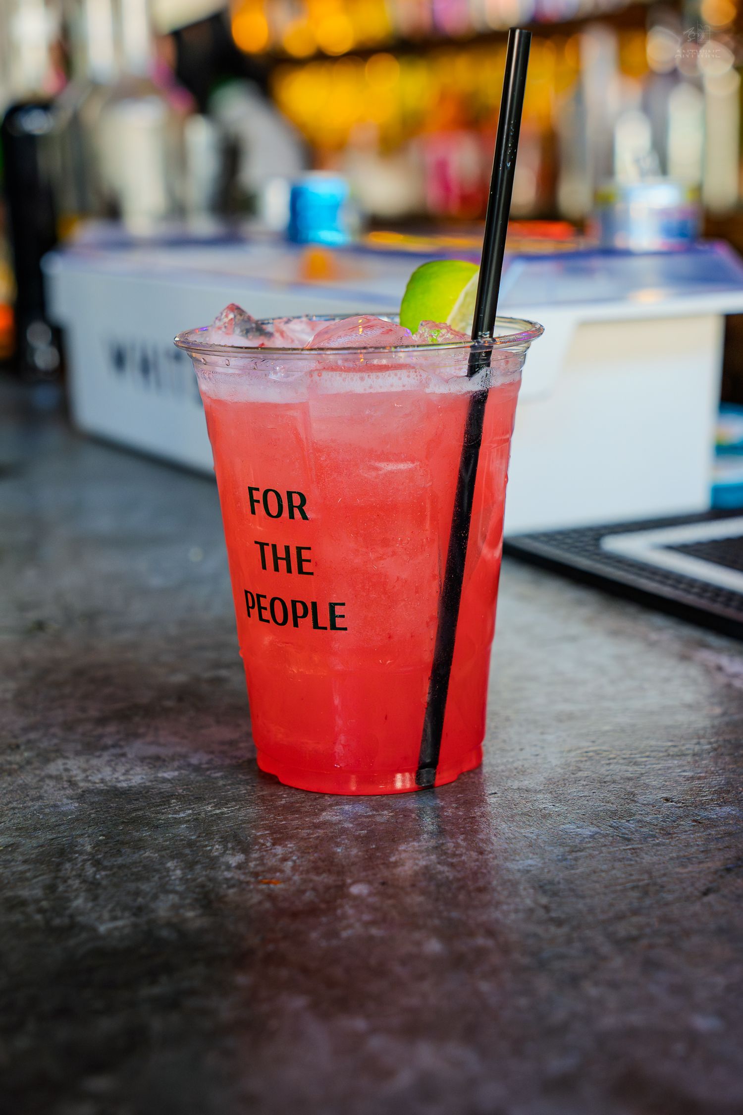 Red cocktail in a plastic cup labeled 'For The People' with ice, lime, and a black straw on a bar counter.