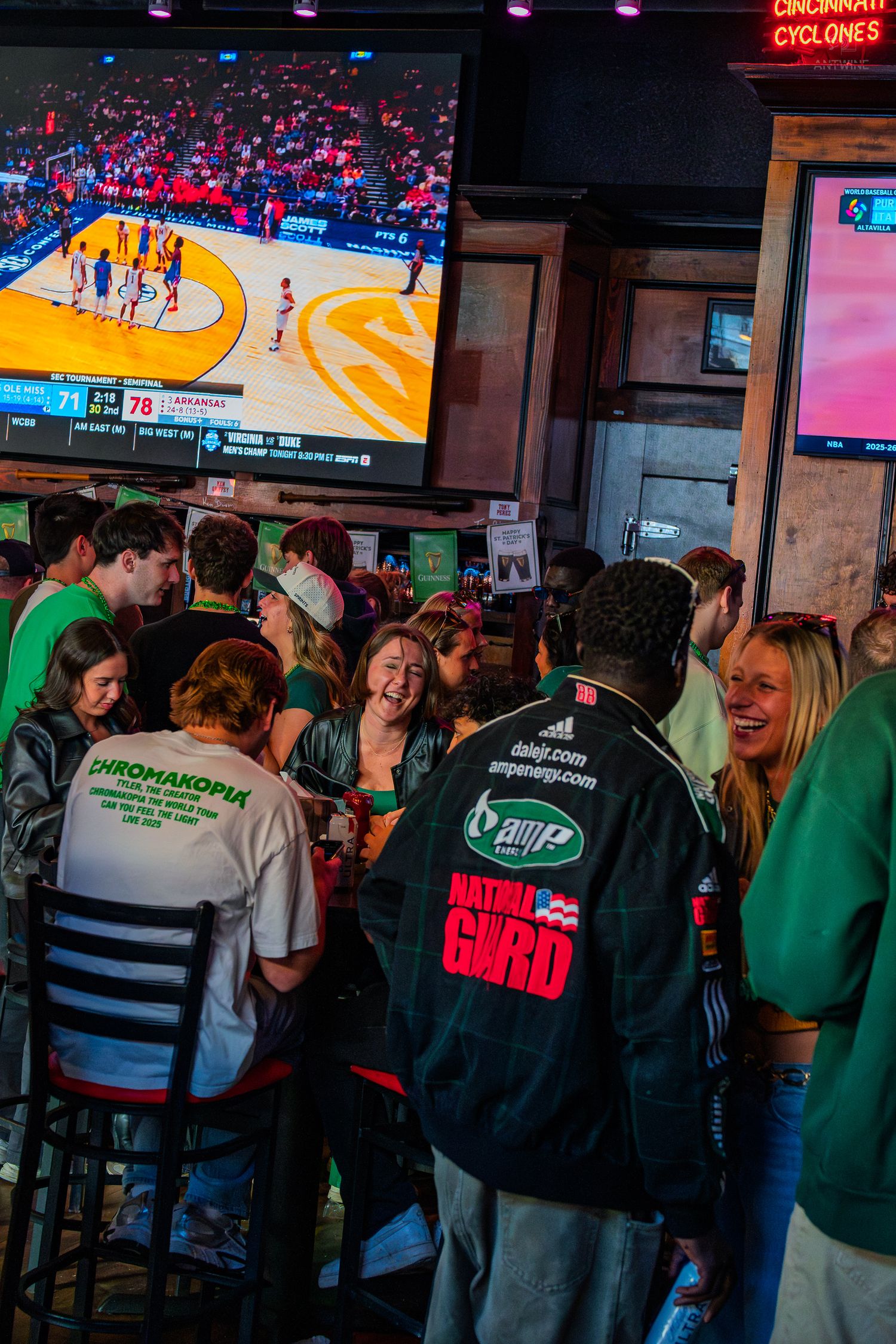 Crowd of young adults watching basketball on TV screens at a lively sports bar, one wearing a National Guard jacket.