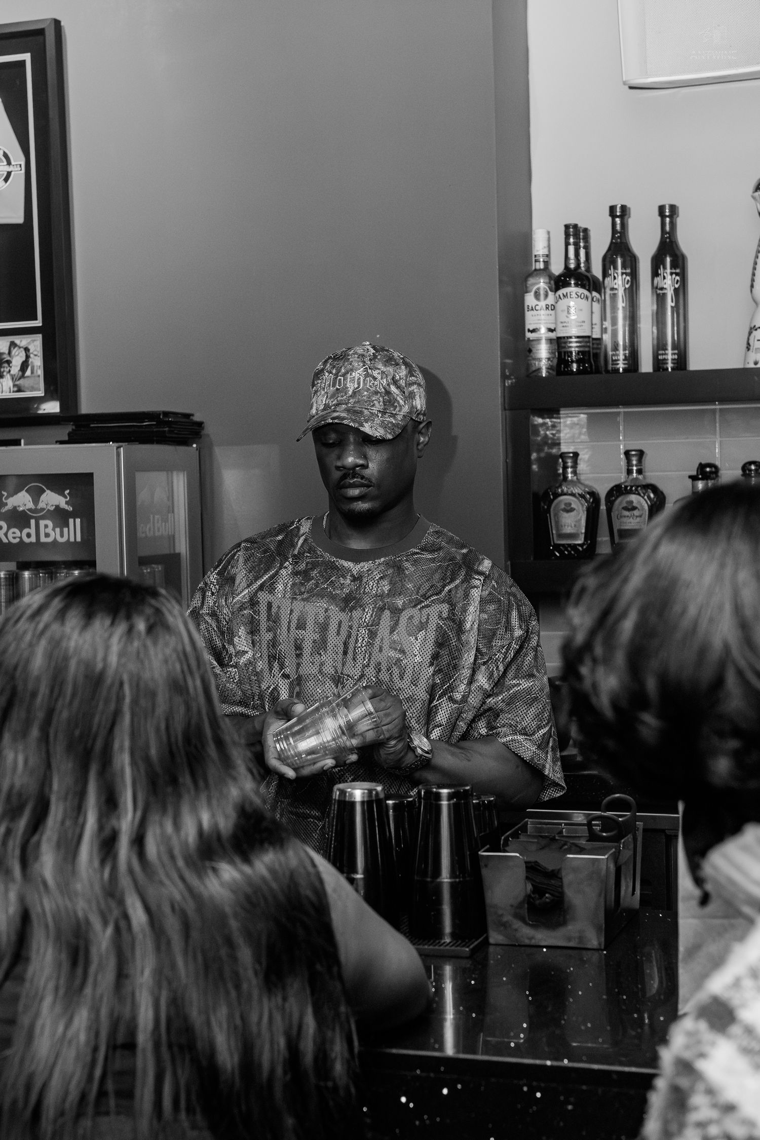 Black and white photo of a bartender in a patterned cap mixing drinks behind a bar with liquor bottles.