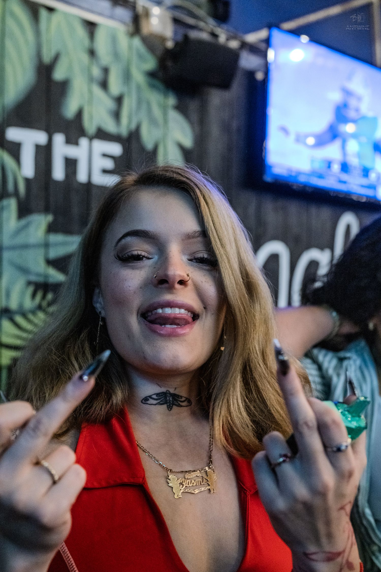 Young woman in red dress sticking out tongue and making hand gestures at a bar event.