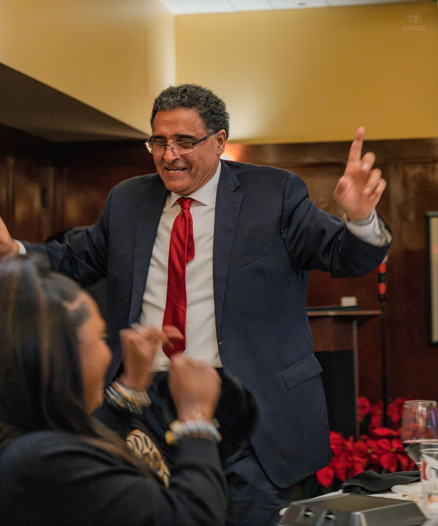 Man in dark suit and red tie gesturing enthusiastically at a formal dinner event.