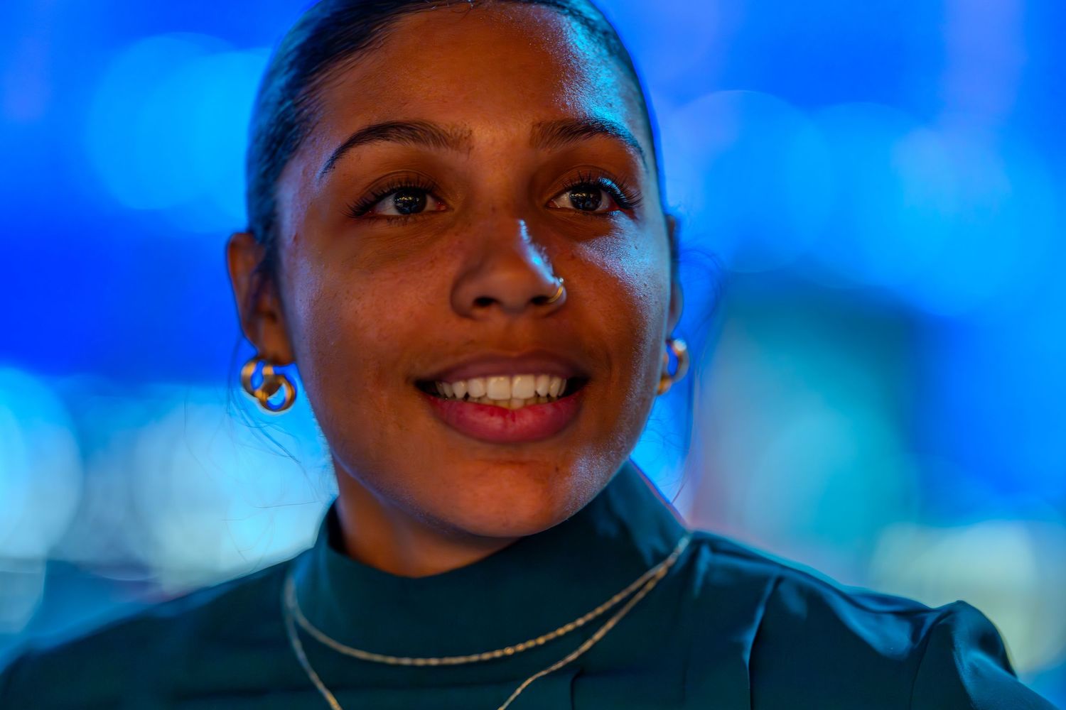 Smiling young woman with hoop earrings and gold necklace against blue bokeh background.