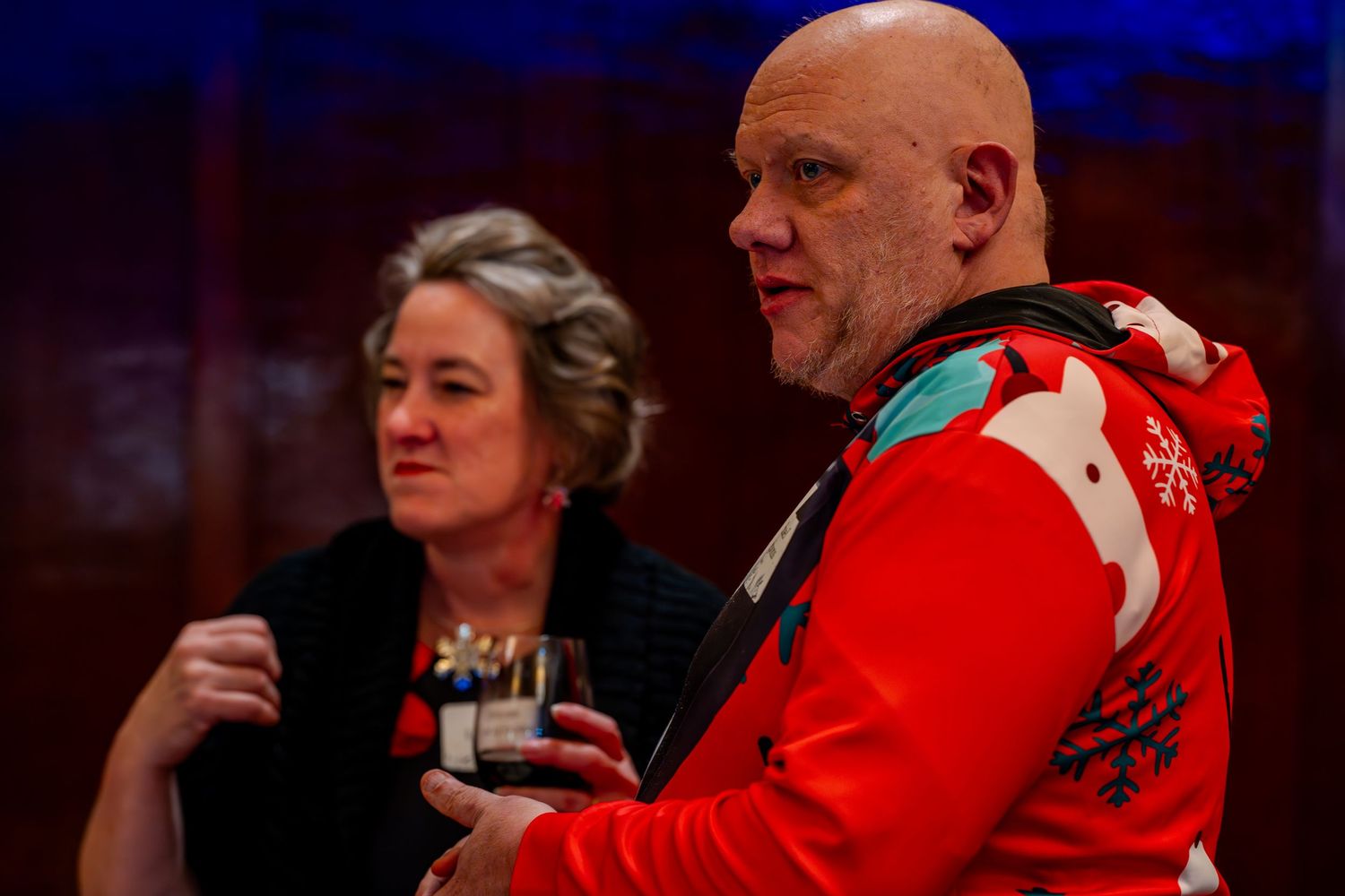 A bald man in a red holiday sweater and a woman in black holding drinks at a festive indoor gathering.