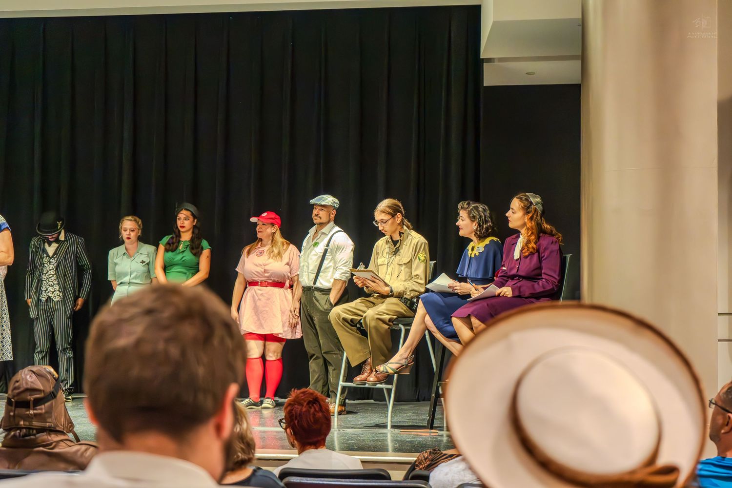 Theater performers in vintage costumes on stage during a live drama production with audience members in the foreground.