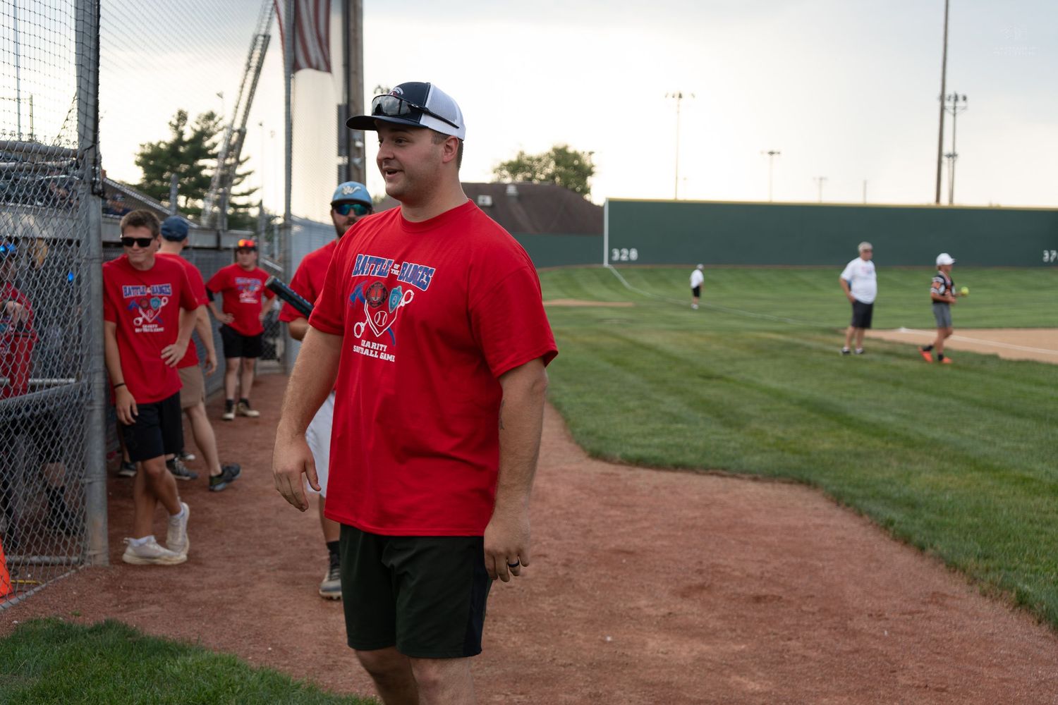 Man in red t-shirt walking along baseball field sideline during outdoor youth baseball event on sunny day.
