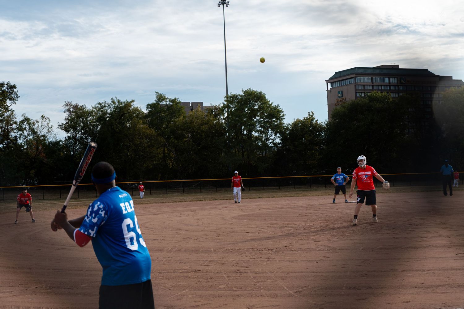 A softball batter in a blue jersey hits a ball into the air as outfielders prepare on a dirt field.
