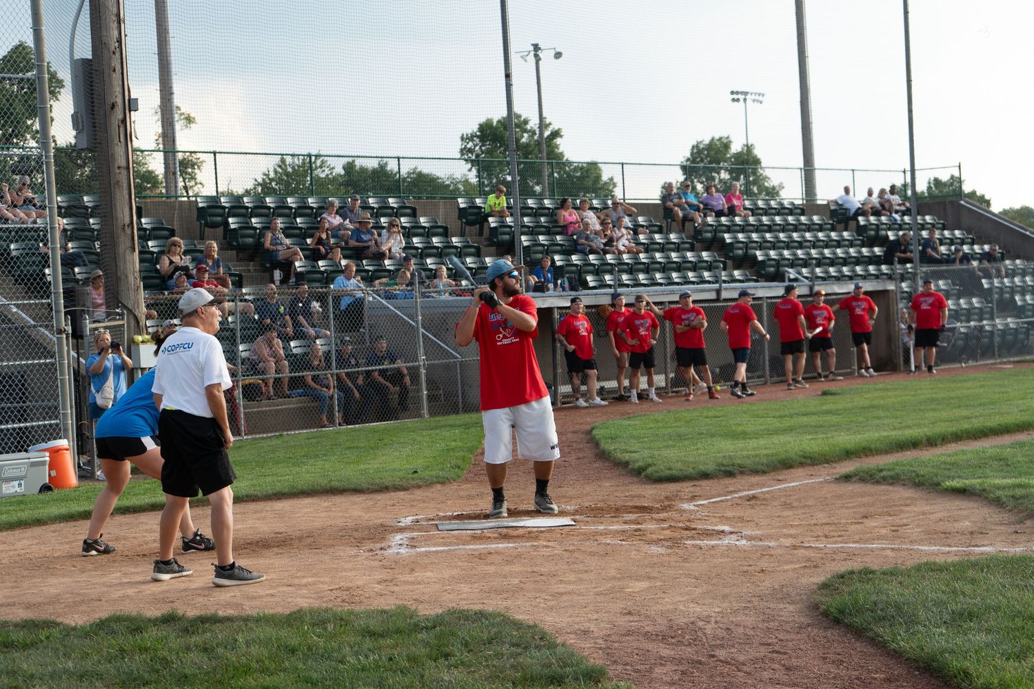 Players line up along the baseline at a softball game in a stadium with green bleachers filled with spectators.