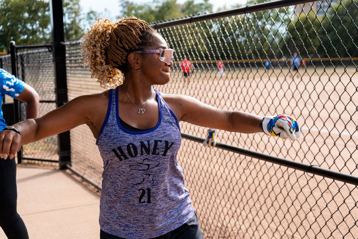 Woman in Honey 21 tank top smiling and stretching by a chain-link fence at an outdoor softball field.