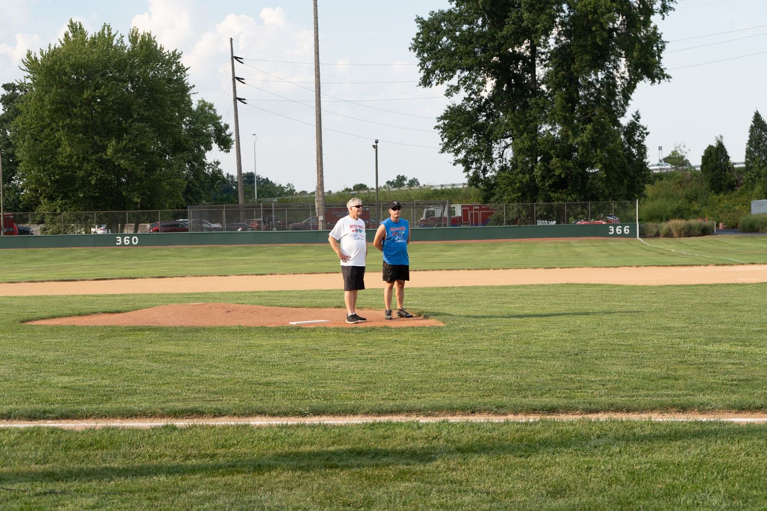 Two kids standing on a baseball pitcher's mound on a sunny day at a green grass field.