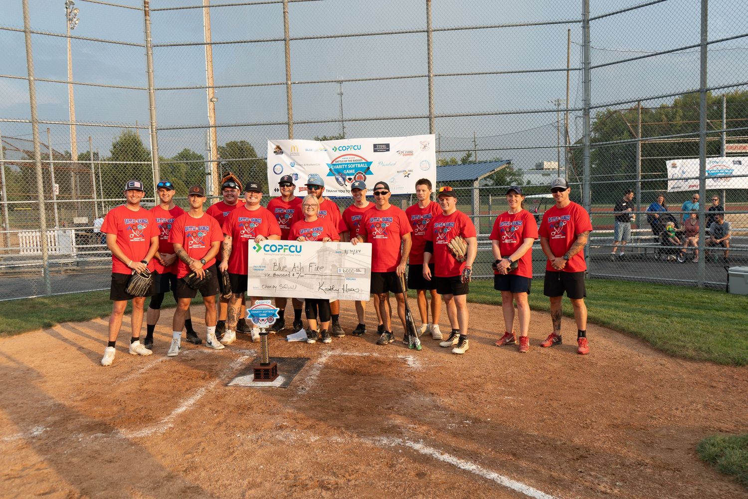 Softball team poses with oversized check at home plate after championship win on outdoor diamond.