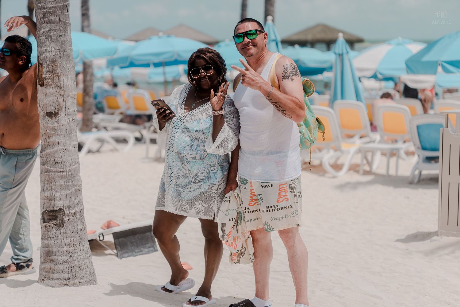 Happy couple posing together on a sunny tropical beach with blue lounge chairs and palm trees in the background.