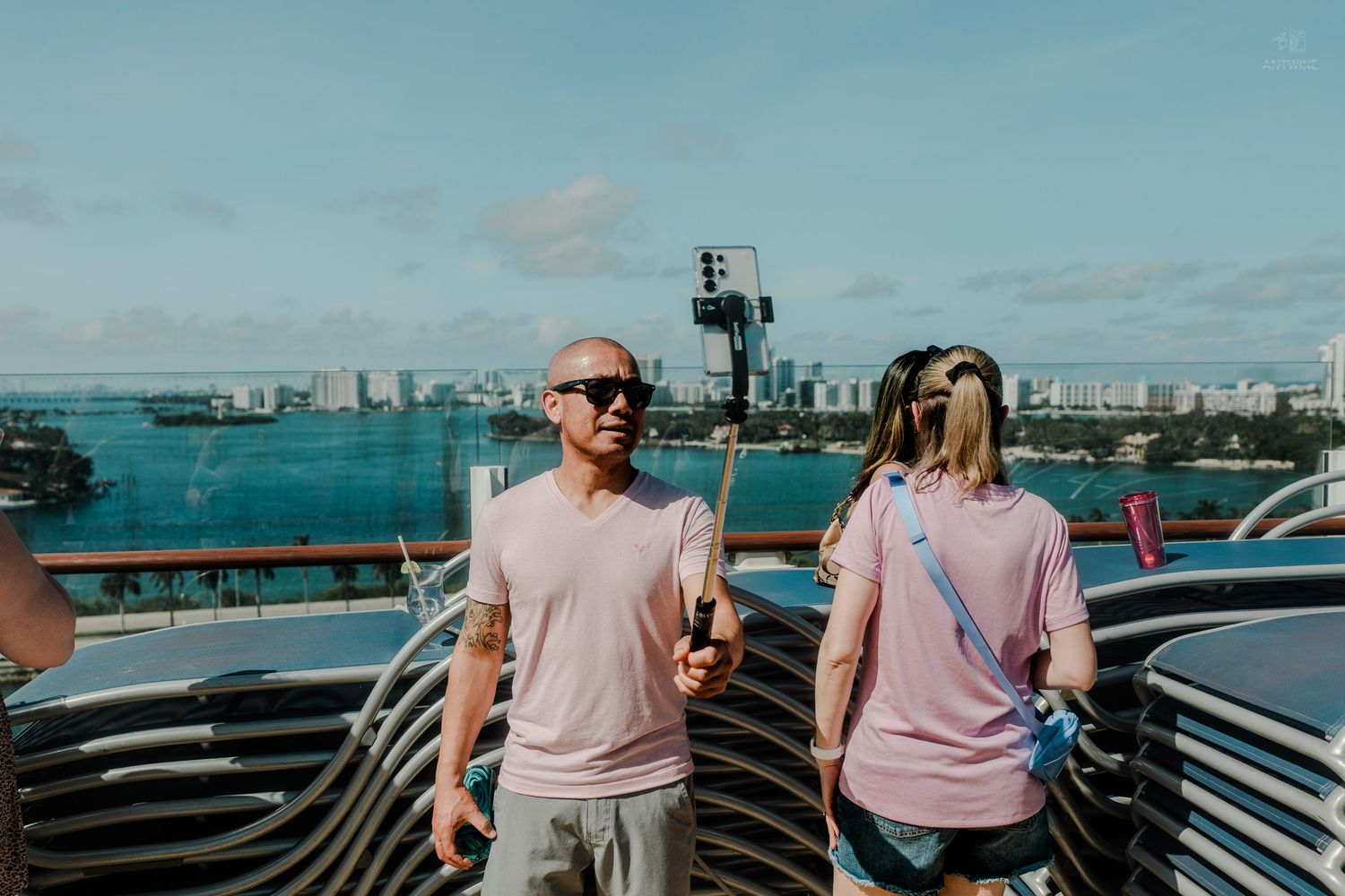 Bald man in pink shirt holding a selfie stick on a cruise ship deck with city skyline behind.