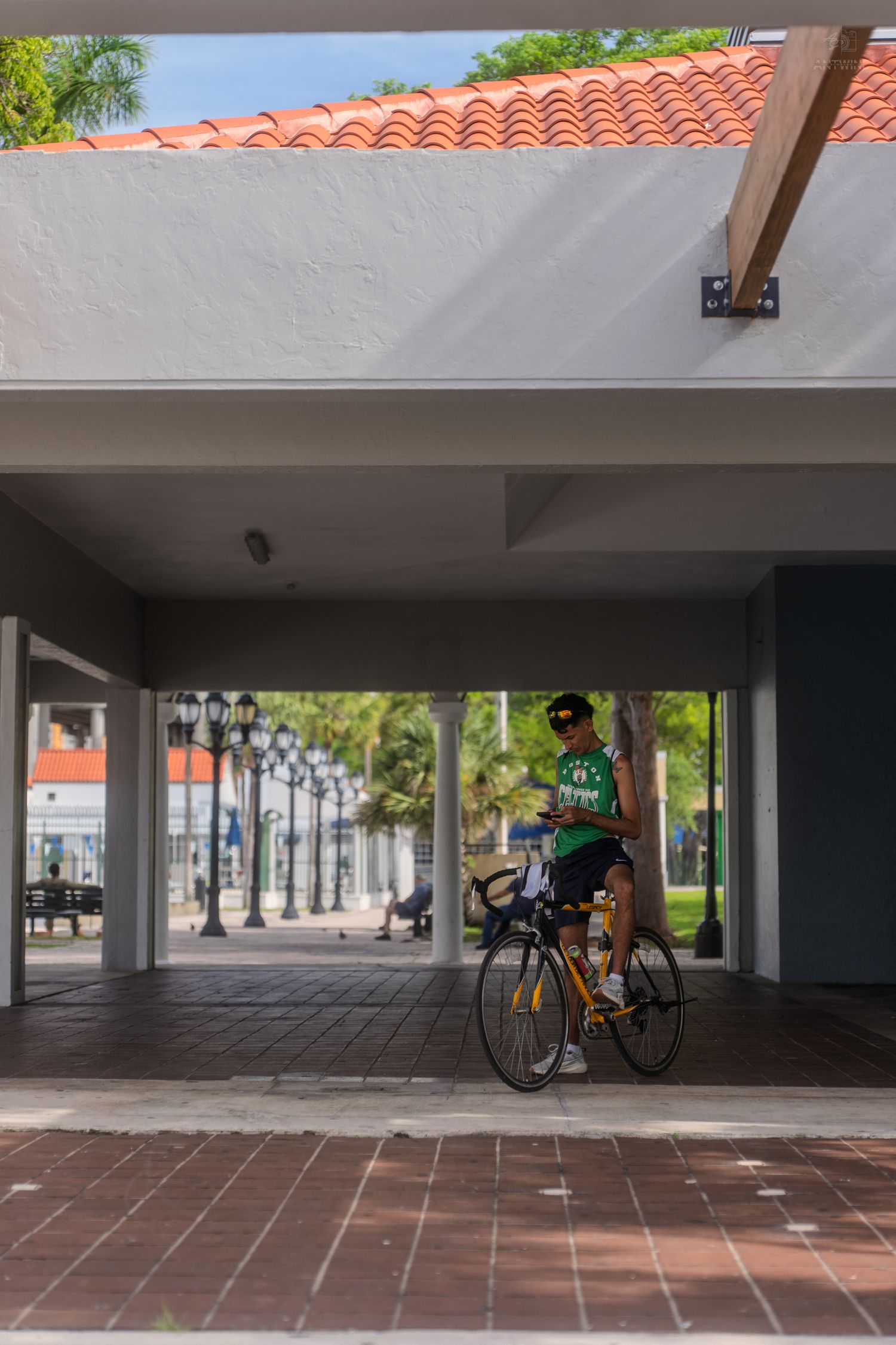 A cyclist in a green jersey pauses under a covered walkway with decorative lamp posts nearby.