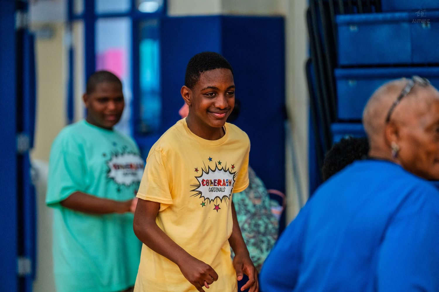 Smiling young man in yellow graphic t-shirt walks indoors with others in colorful shirts in background.