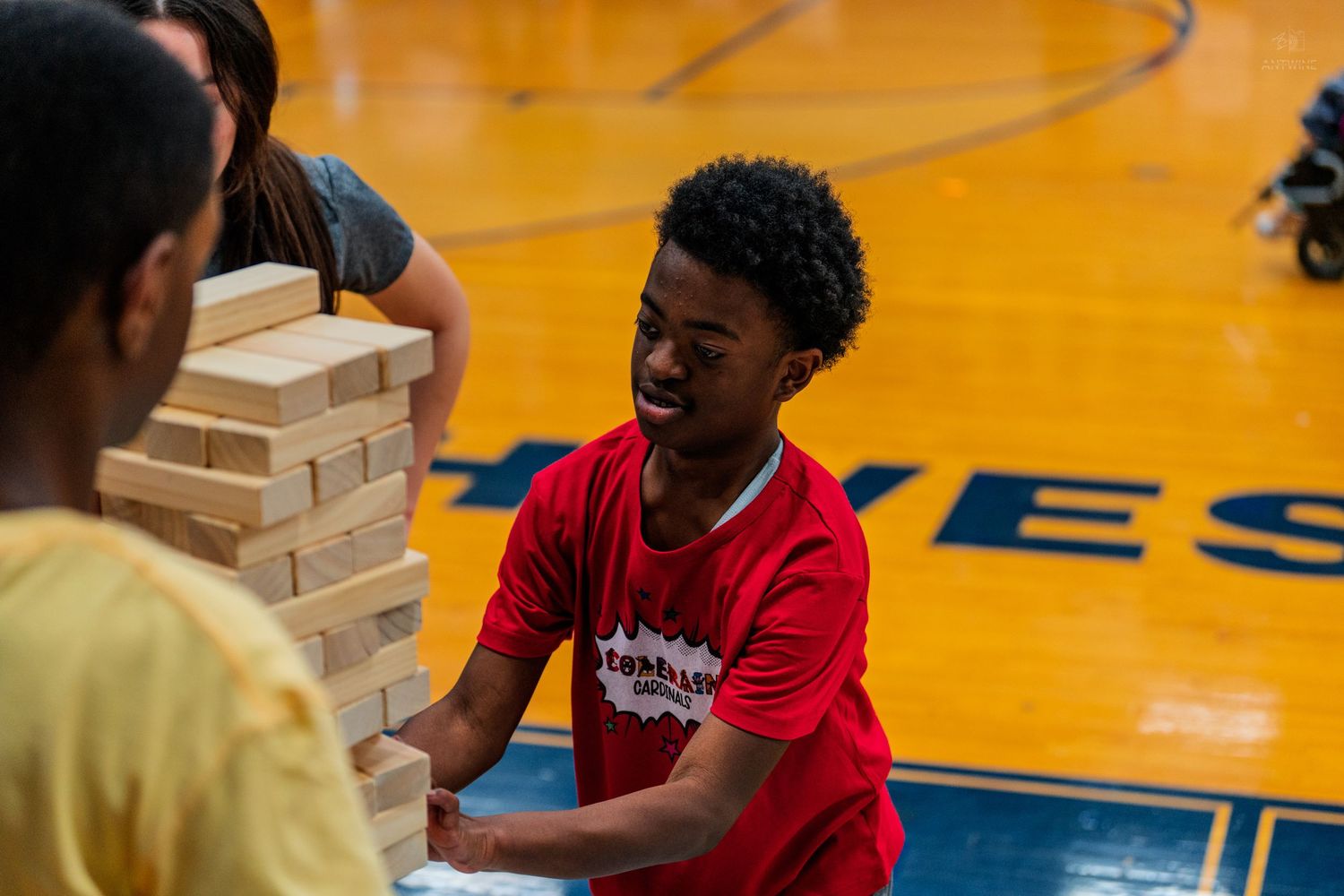 A boy in a red shirt laughs while playing Jenga on a gymnasium floor during a school activity.