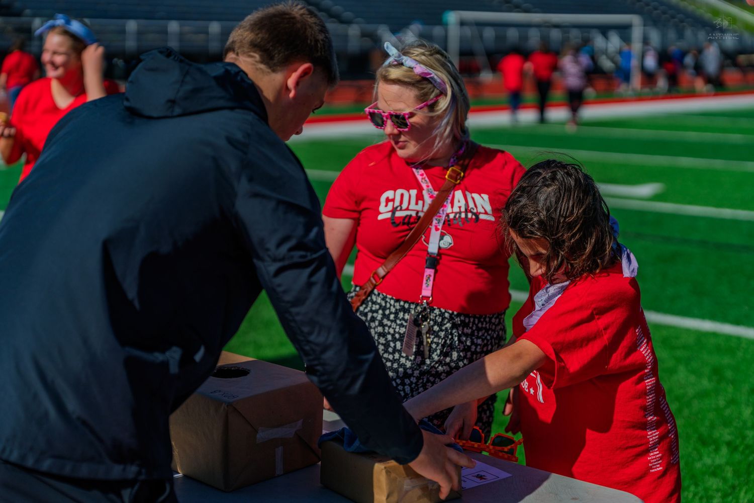 Adults and child in red Colorado shirts opening a box on a table at an outdoor sports field event.