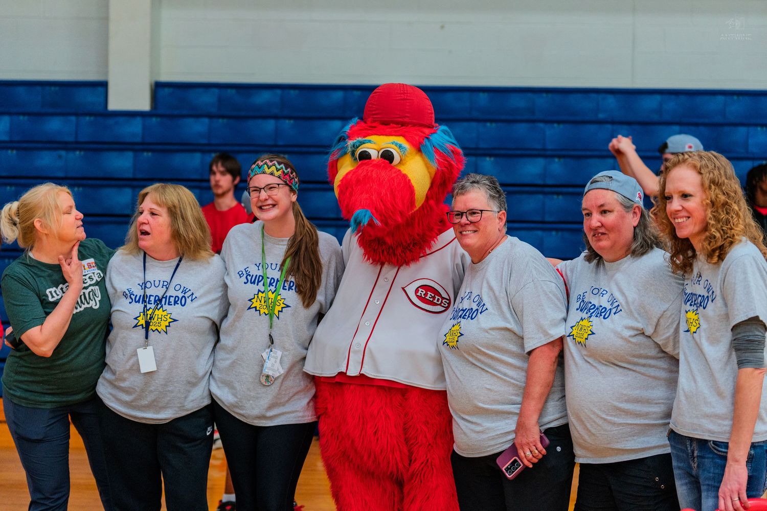 Group of women in matching gray shirts posing with a red bird mascot in a gymnasium.