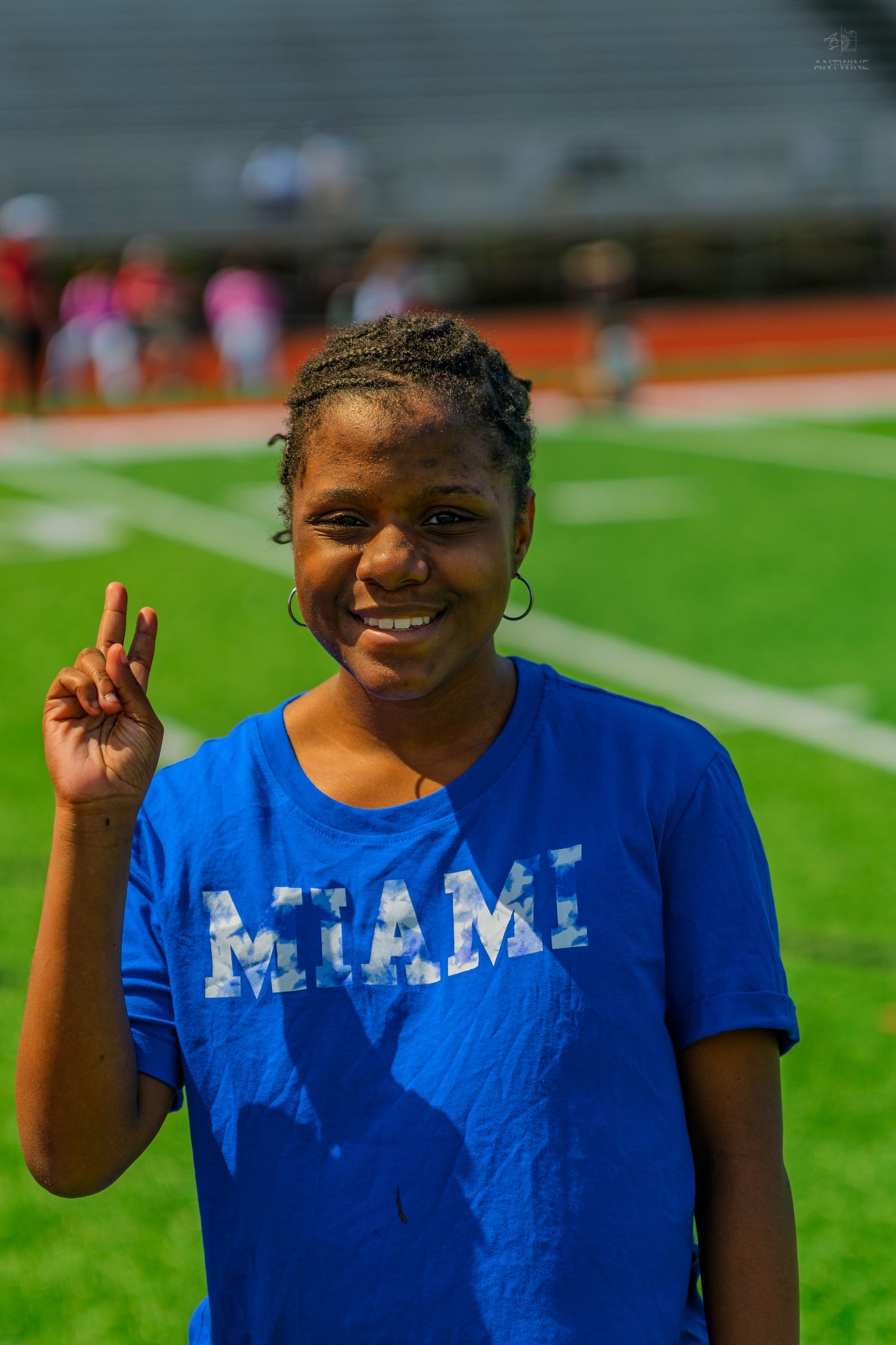 Young woman in blue Miami t-shirt smiling and giving peace sign on green football field.