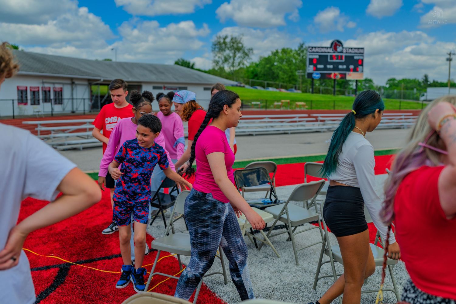 Students walk on red carpet at an outdoor track and field event with folding chairs and a scoreboard visible in background.