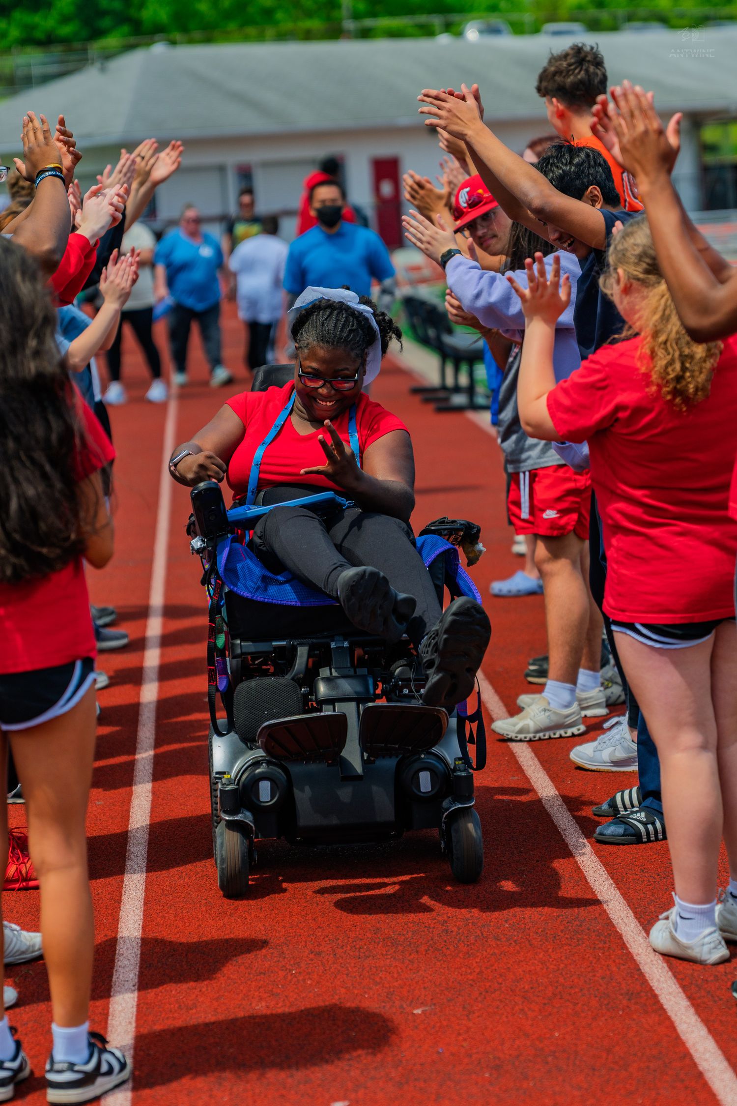 Athlete in power wheelchair receives high-fives from cheering crowd on red track at Special Olympics event.