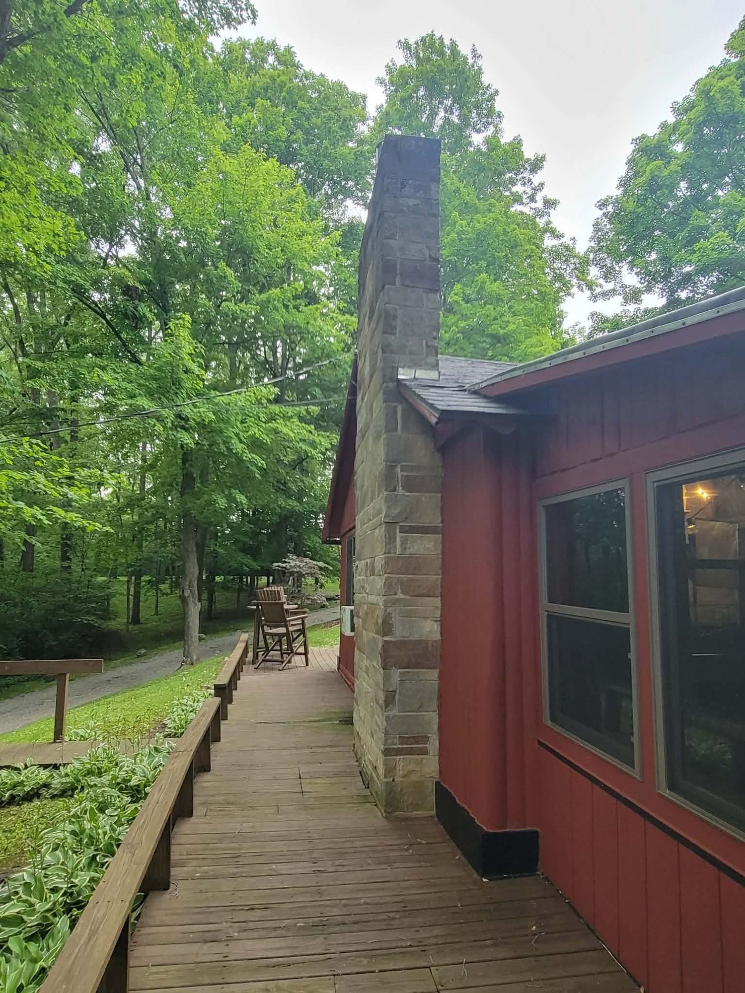 Red cabin with stone chimney on wooden deck surrounded by lush green trees in a wooded setting.