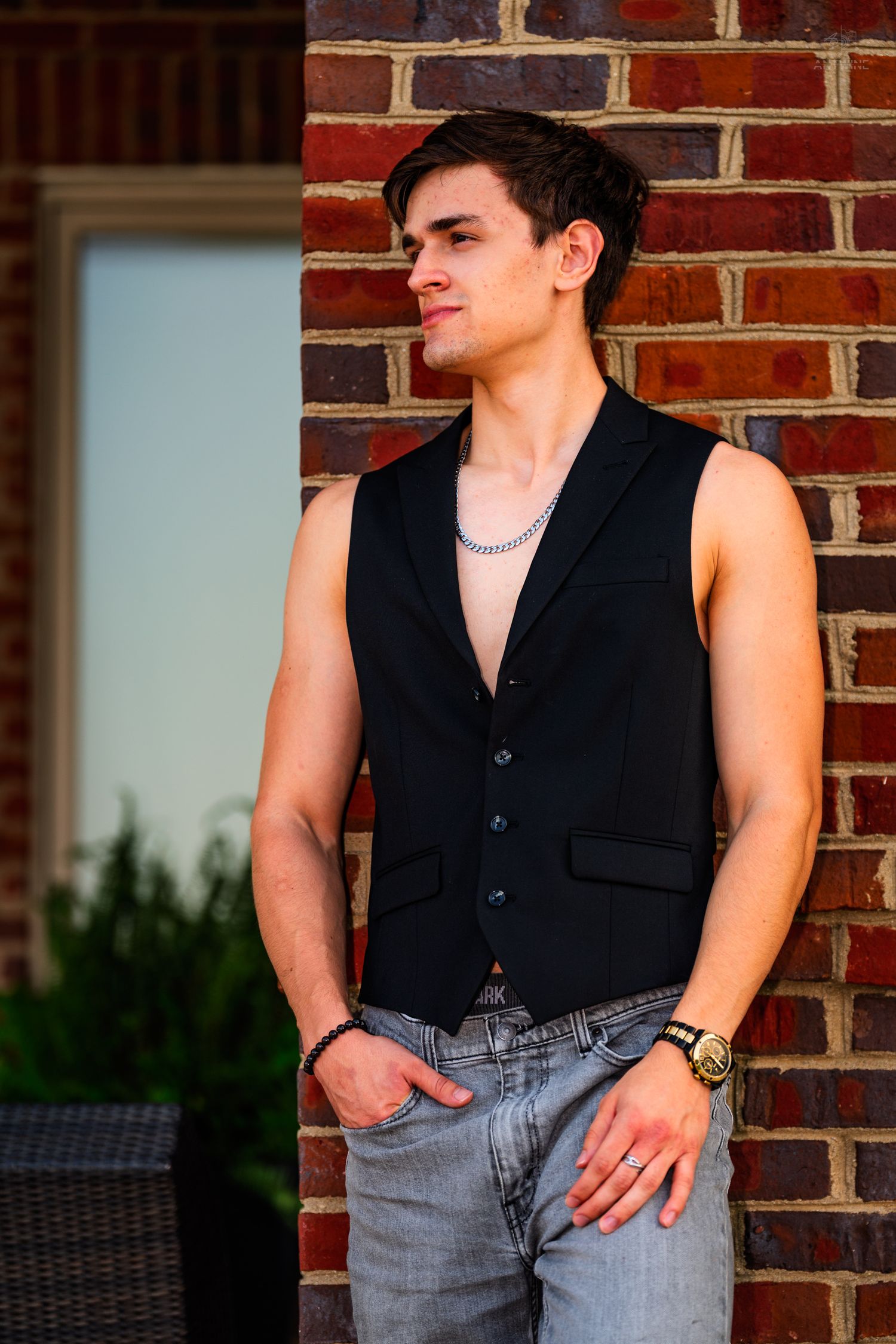 Young man in black vest and jeans with chain necklace poses by brick wall outdoors.