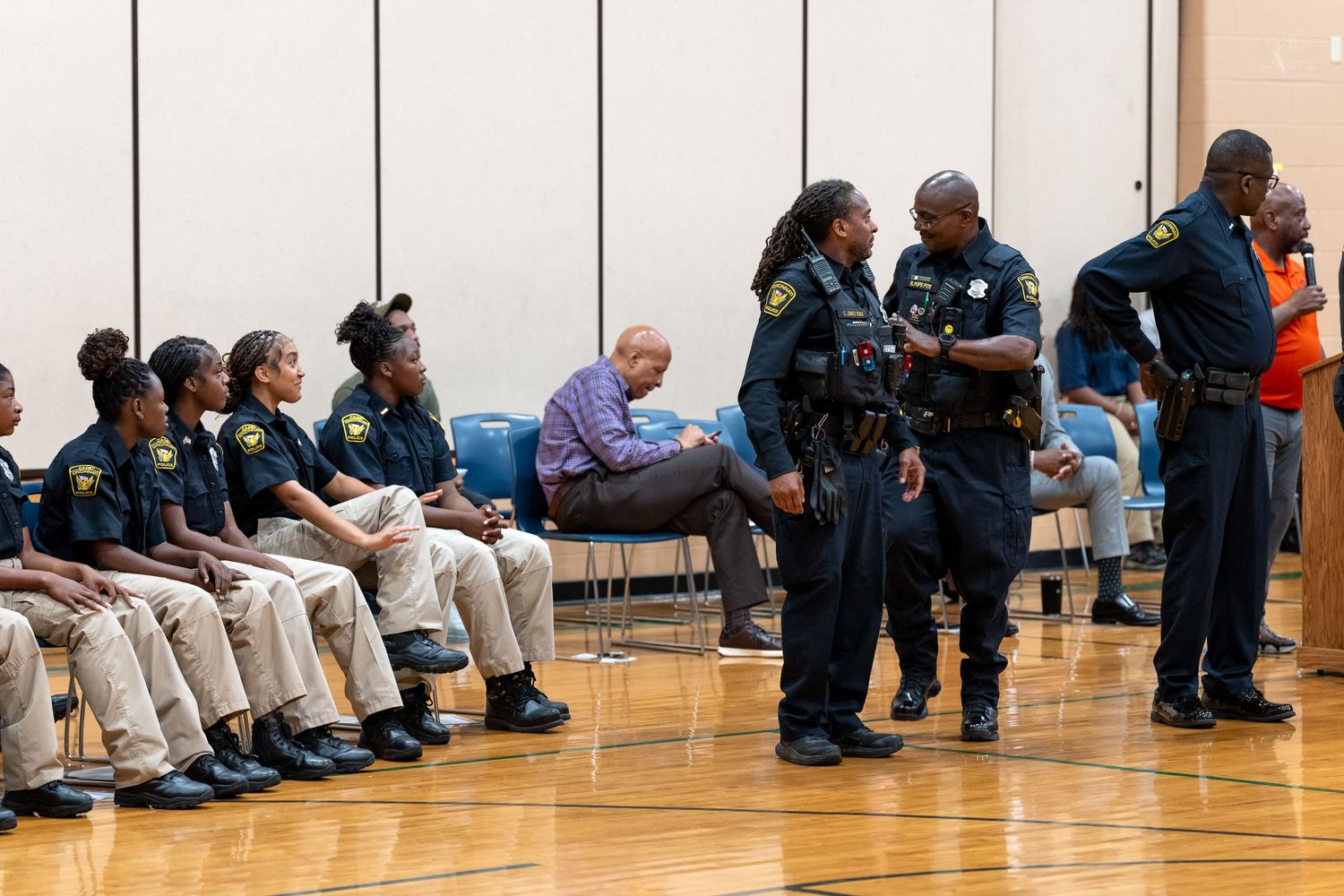 Law enforcement officers in uniform gather in a gymnasium, with several seated along the wall during a training event.