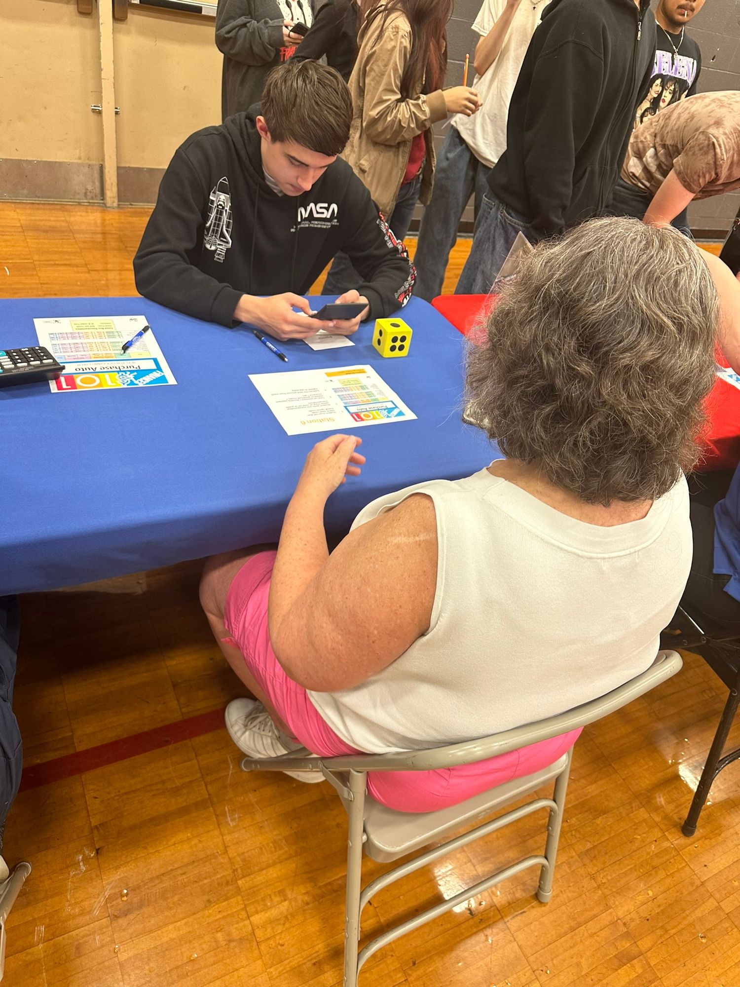 Young man uses a yellow die while helping an older woman at a blue table during an educational fair.