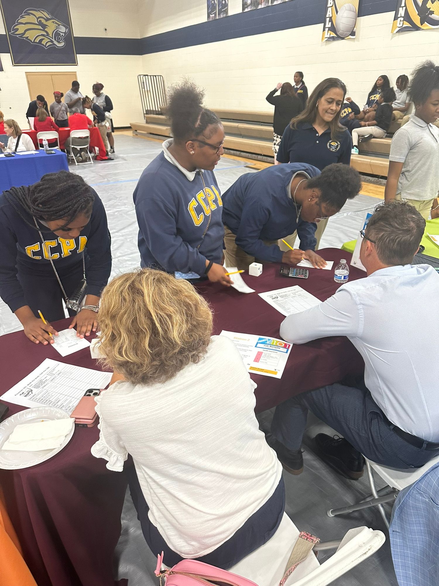 Students in CCPA uniforms collaborate with adults at a maroon table during a school event in a gymnasium.
