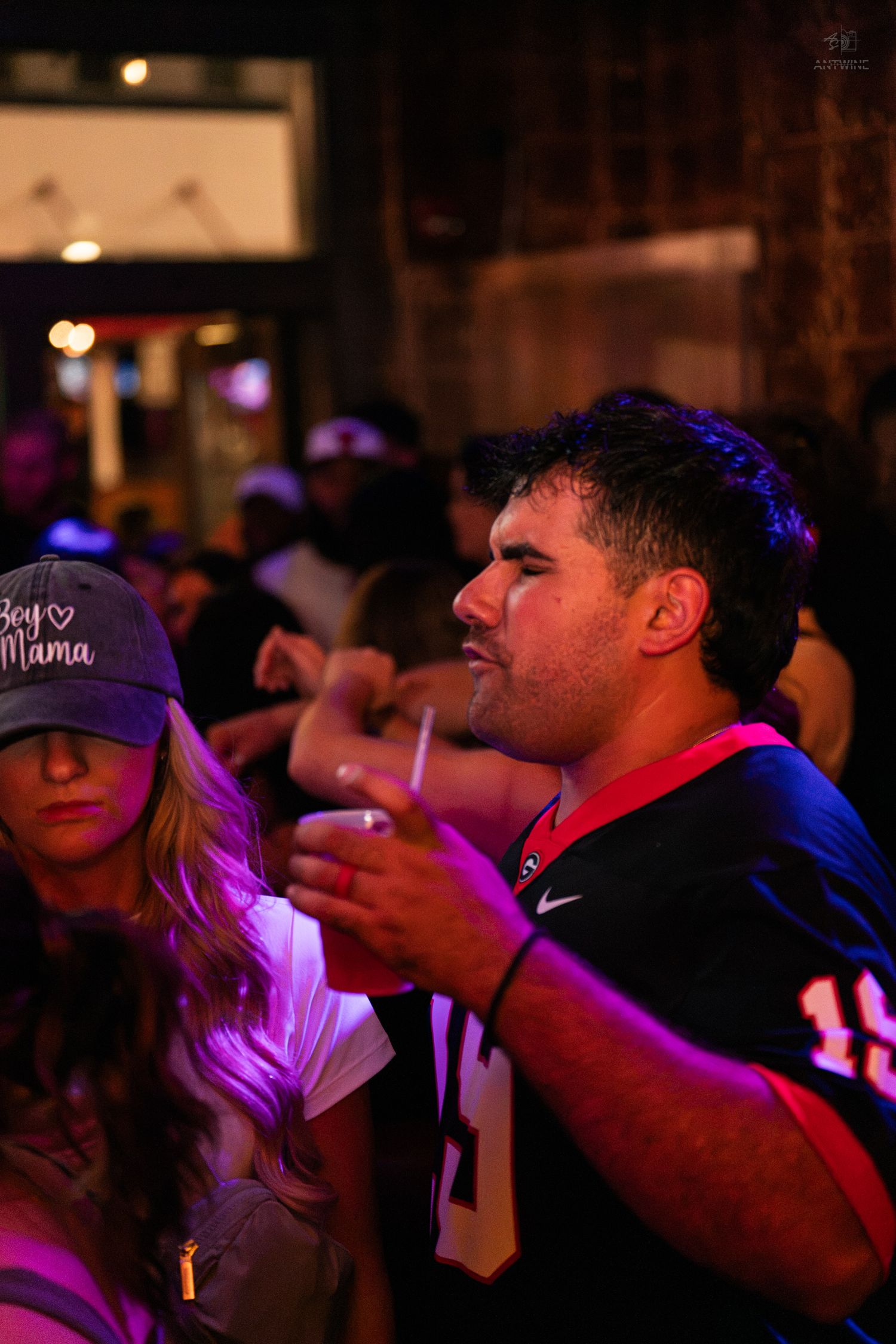 Man in football jersey holds drink at crowded nightclub event with purple lighting.