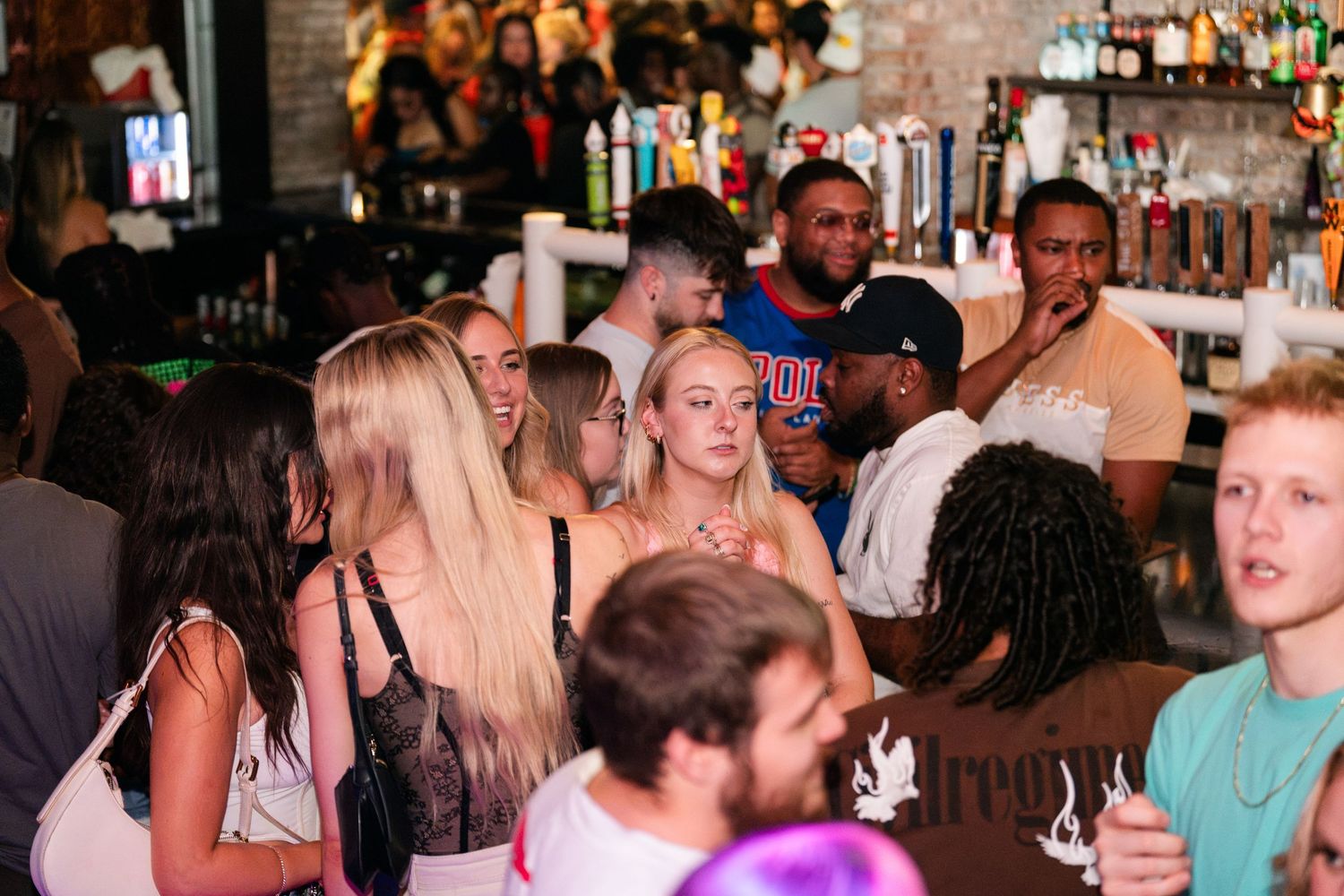 Diverse crowd of young people mingling and socializing inside a busy bar or nightclub.