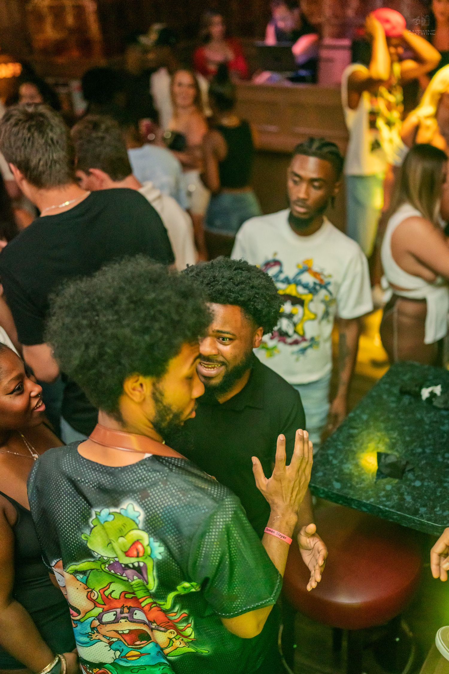 Two men engaged in lively conversation at a crowded nightclub with warm ambient lighting.