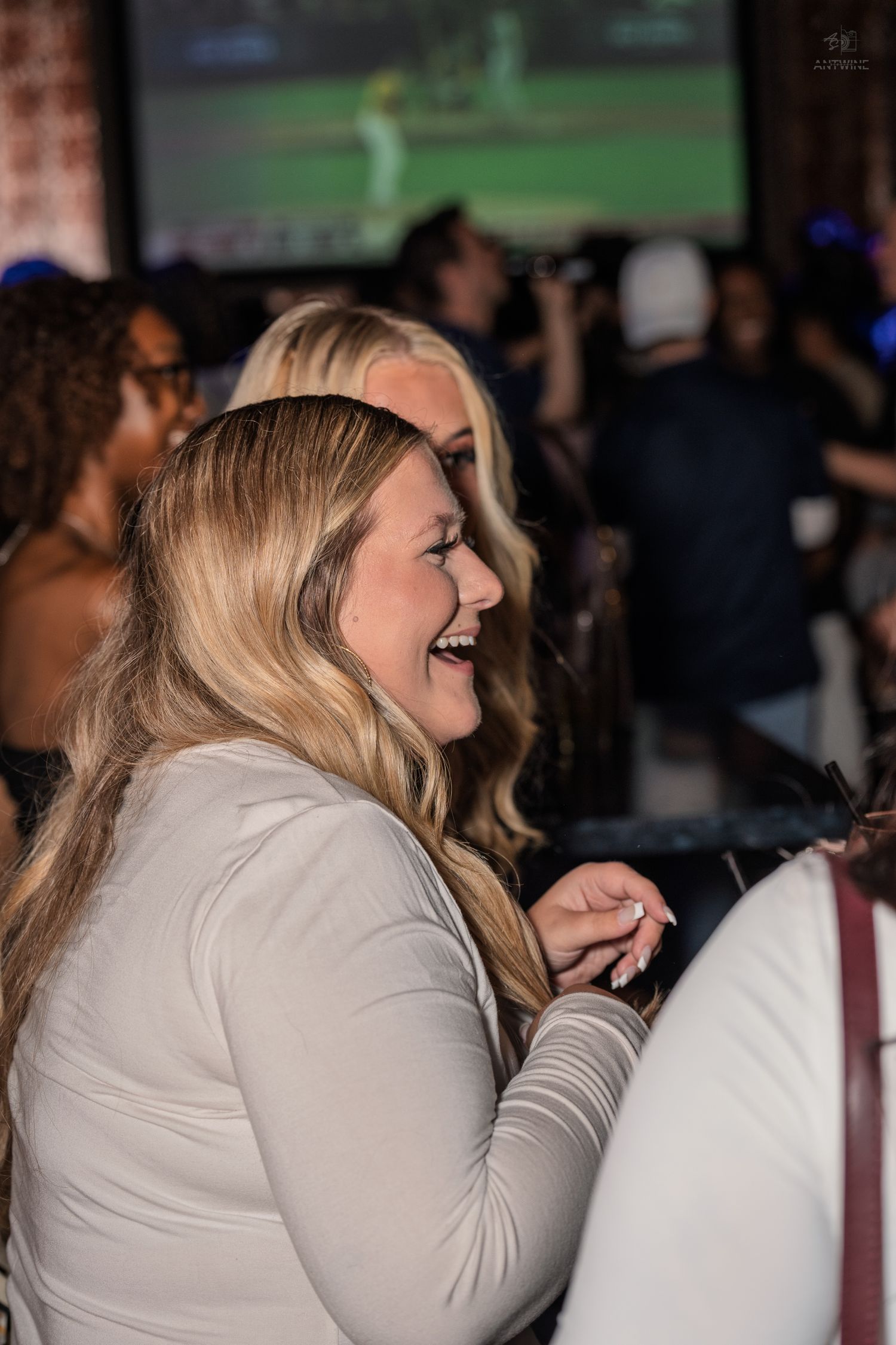 Laughing blonde woman in white striped top enjoys herself at a crowded sports bar with TV screen.