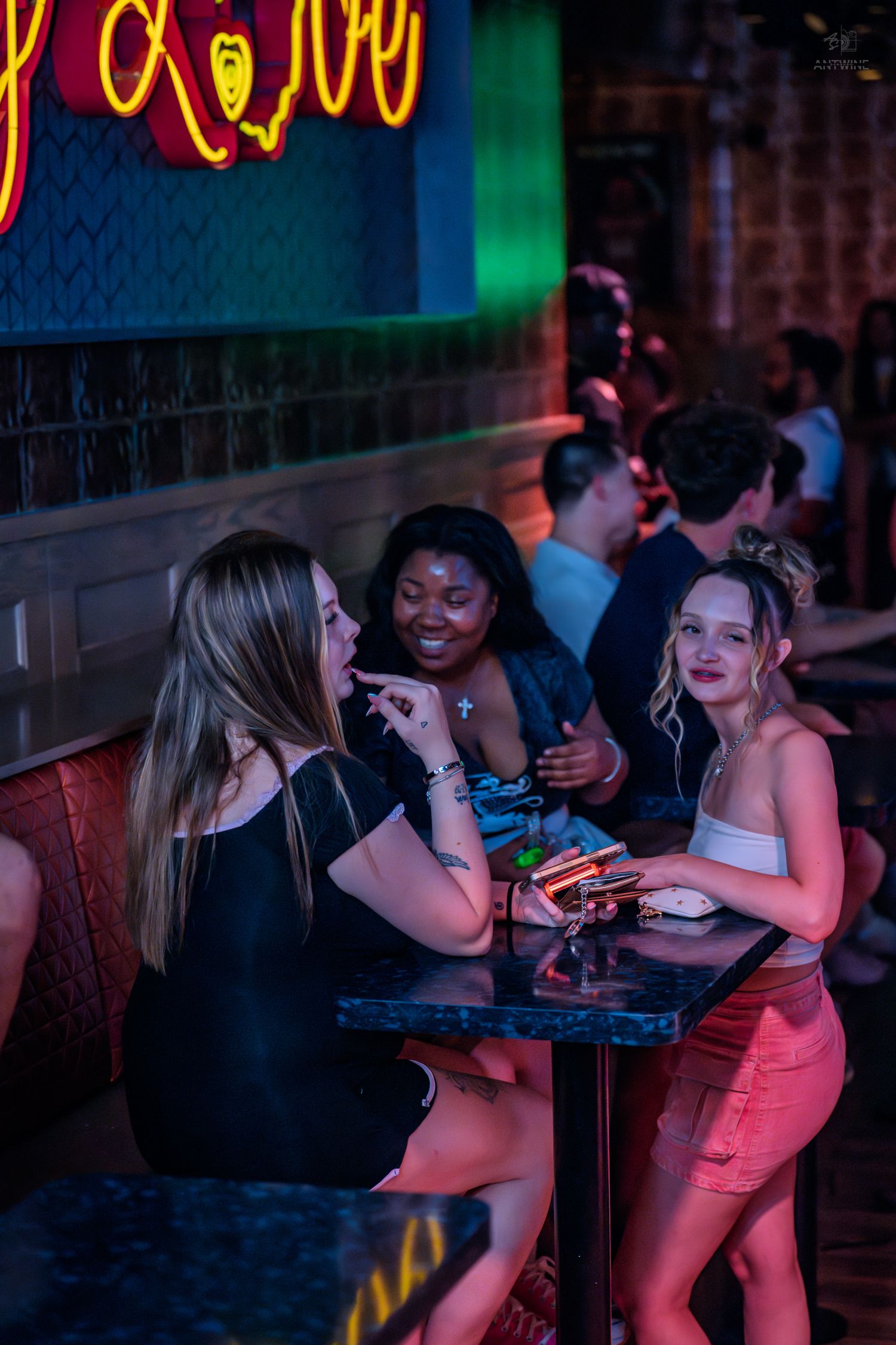 Three women smile and chat together at a bar table at night.