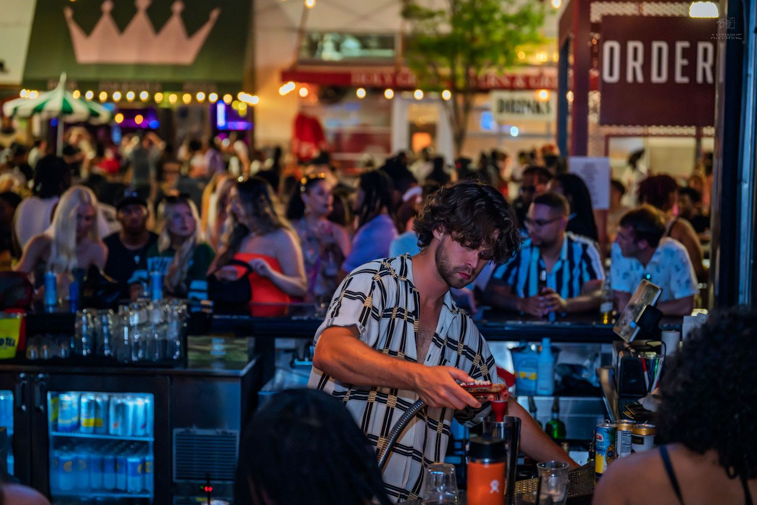 Bartender in patterned shirt mixing drinks at a busy outdoor bar with crowd behind.
