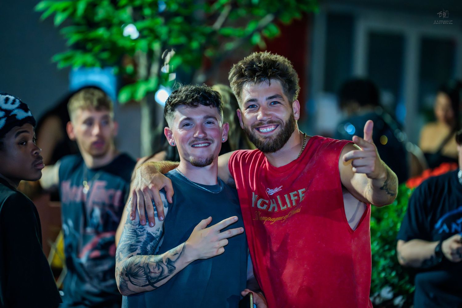 Two smiling young men pose together at a nighttime outdoor event, one giving a thumbs up.