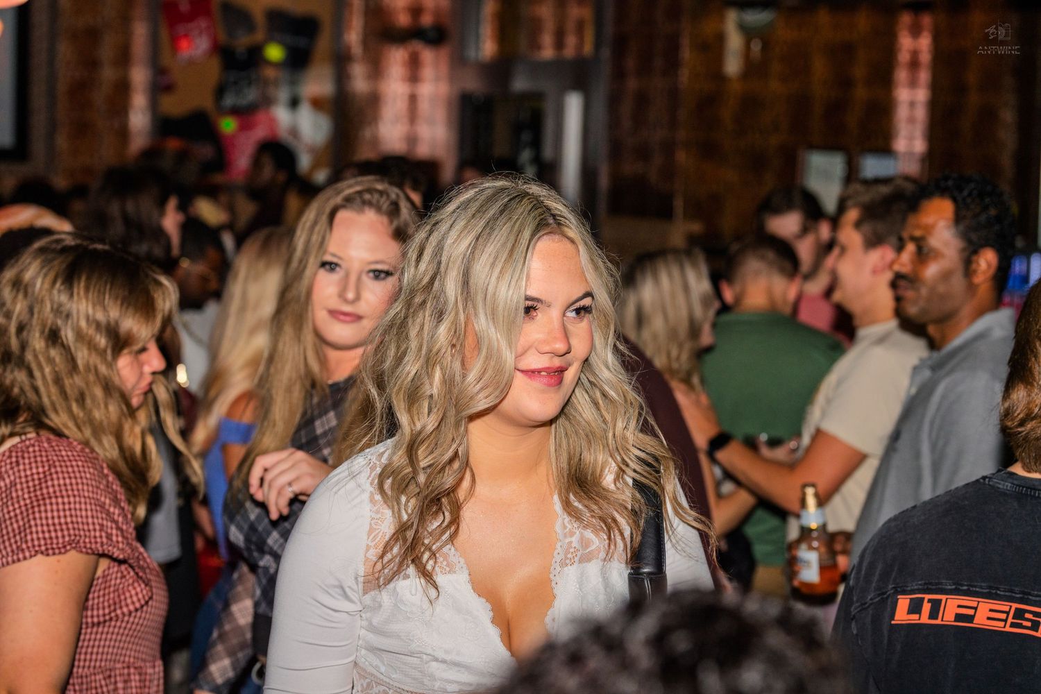 Two blonde women smiling and socializing in a busy bar or nightclub crowd at night.