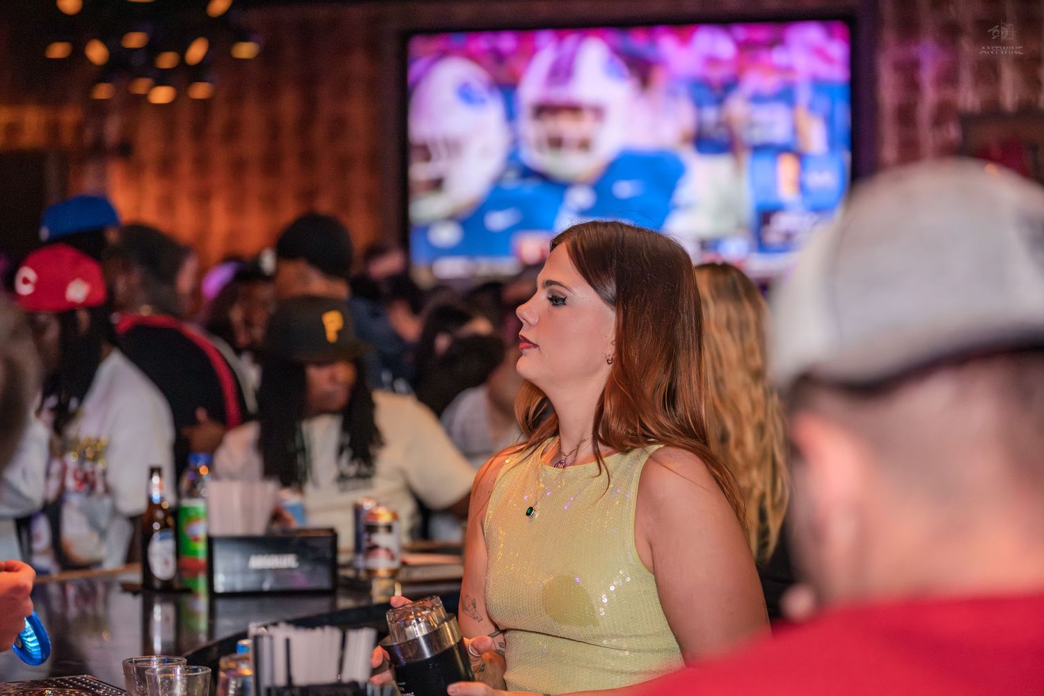Woman in yellow top holding a drink at a lively sports bar with football game on screen.