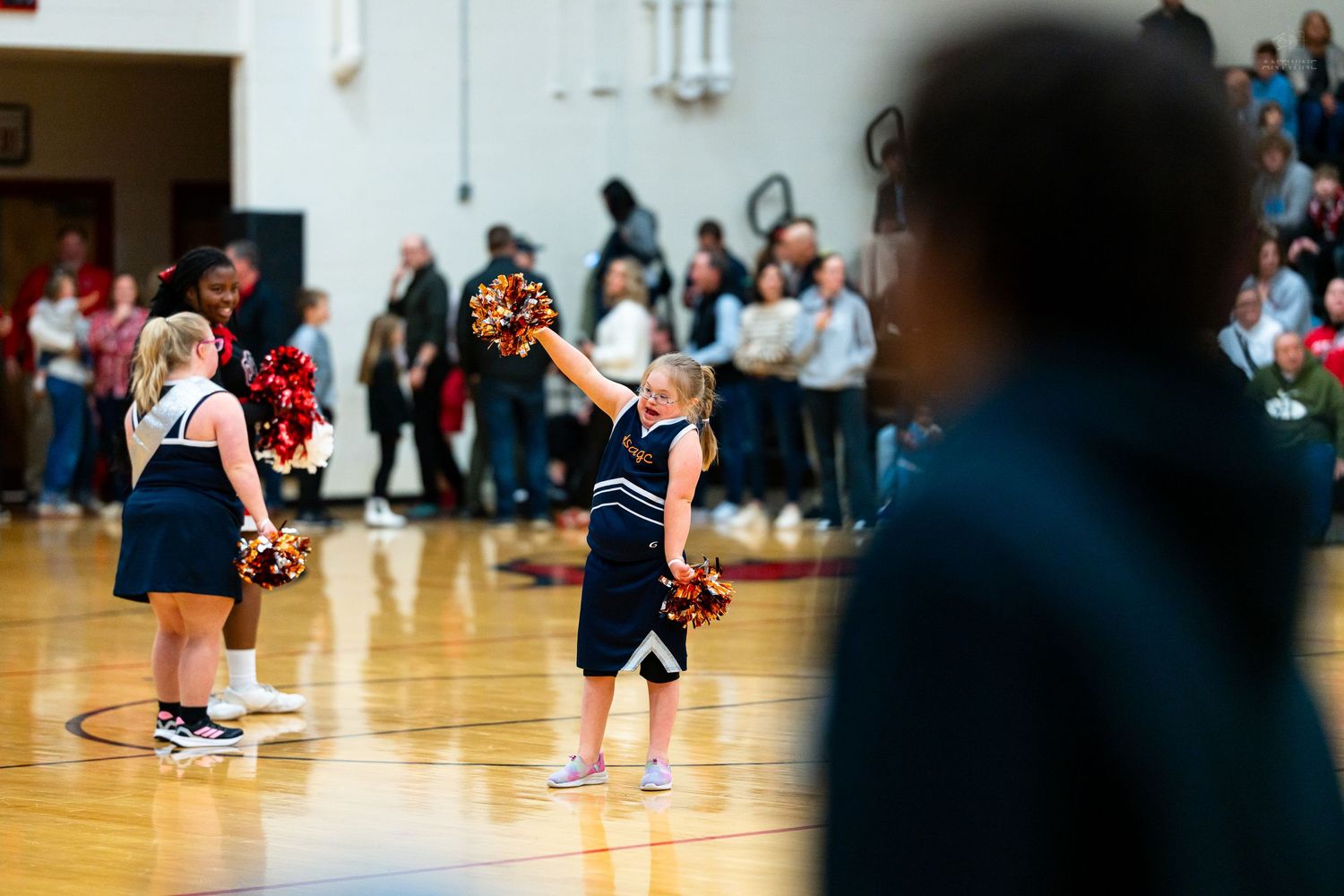 Young cheerleaders in navy uniforms perform with pom-poms on basketball court before a large crowd.