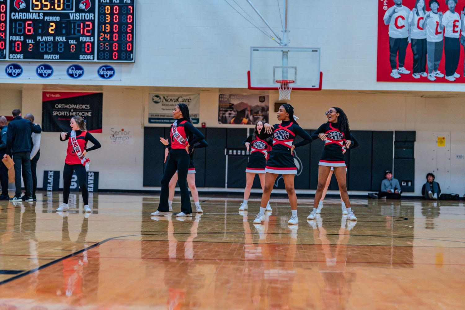 High school cheerleaders in red and black uniforms perform a synchronized dance routine on a basketball court.