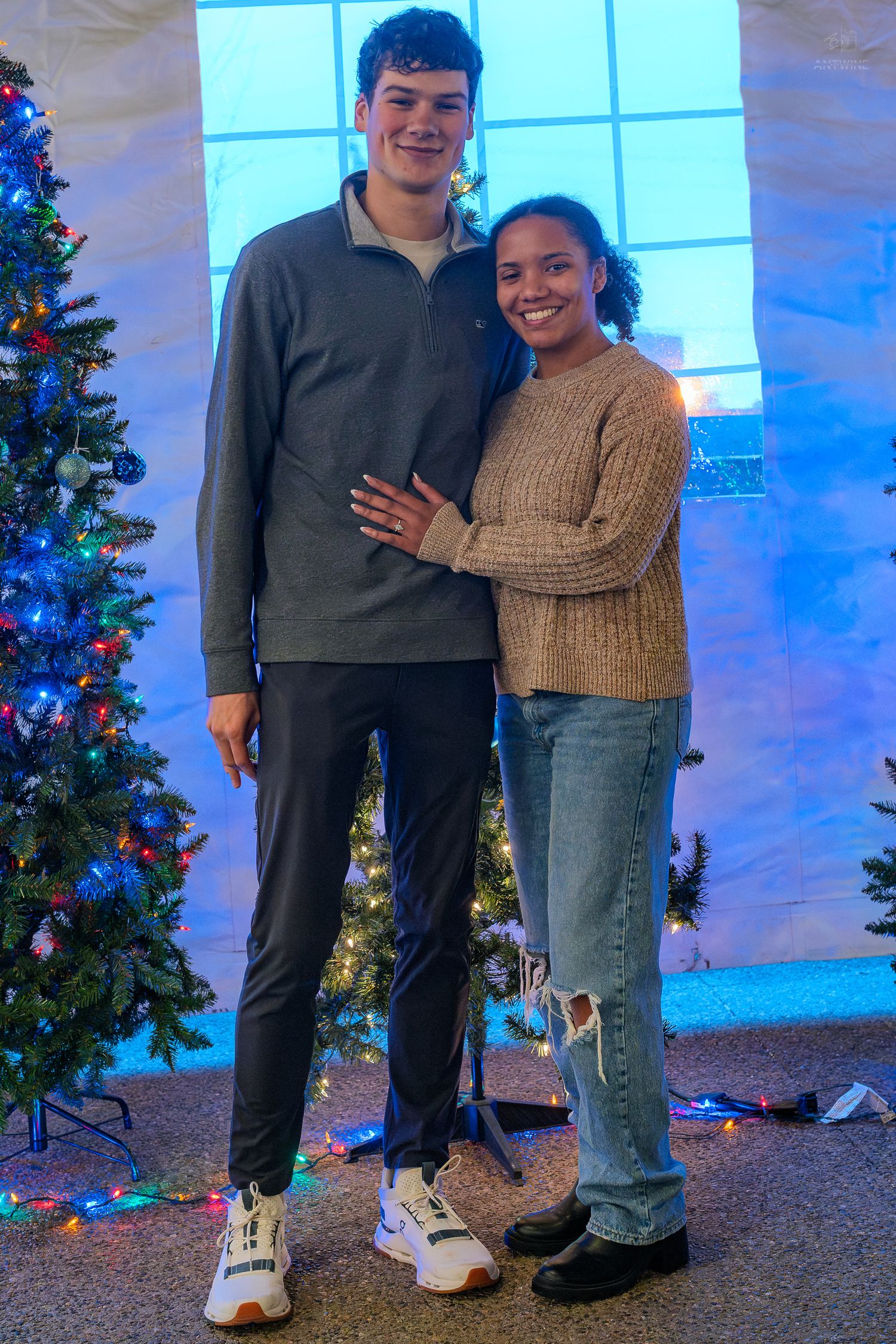 Tall young couple posing together in front of a decorated Christmas tree at a holiday party.