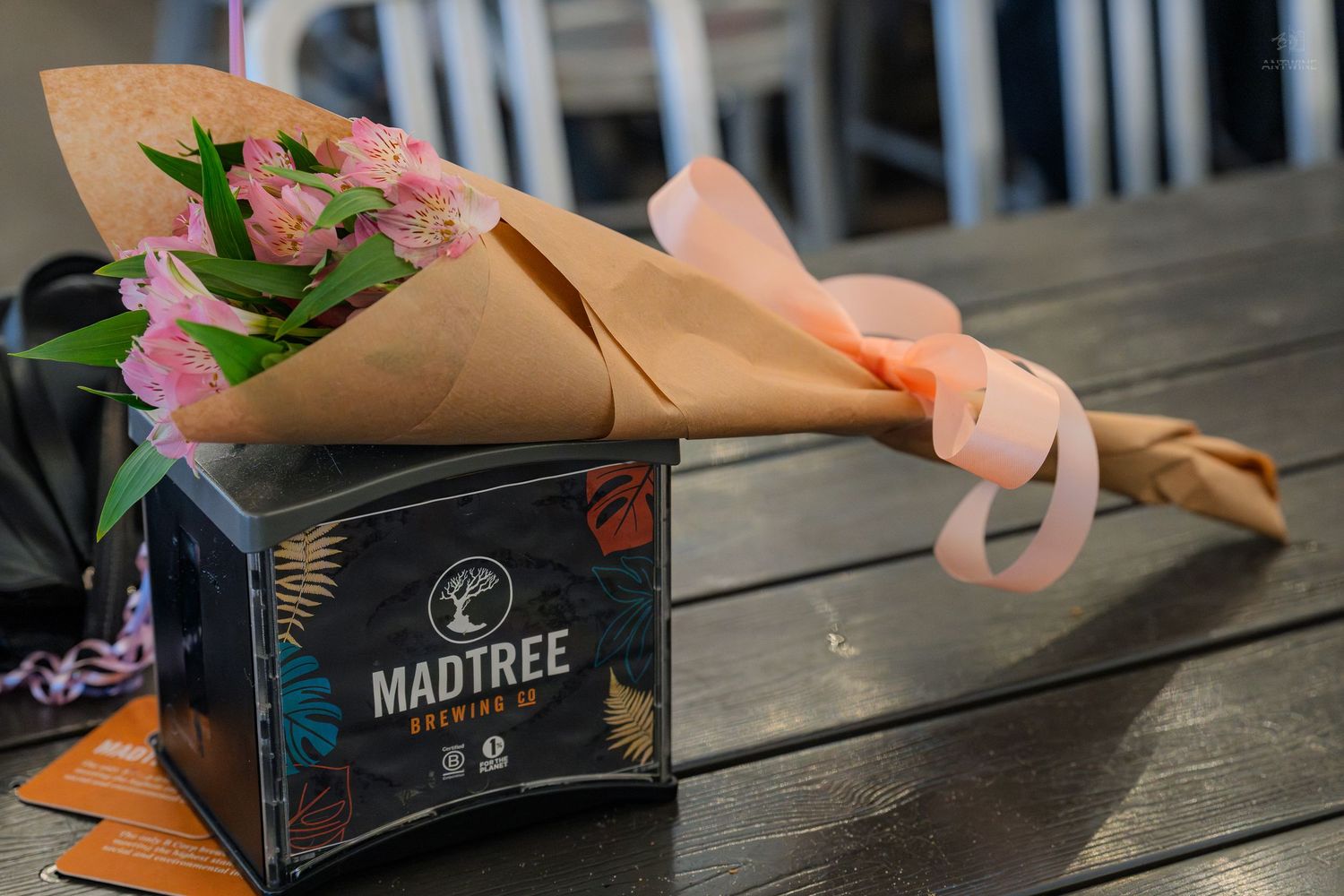 A bouquet of pink roses wrapped in kraft paper rests atop a MadTree Brewing Co. box on a wooden table.