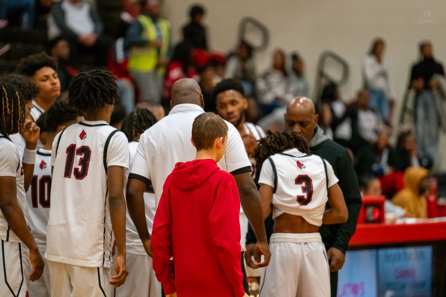 High school basketball team gathers in sideline huddle with coaches during game timeout in packed gymnasium.