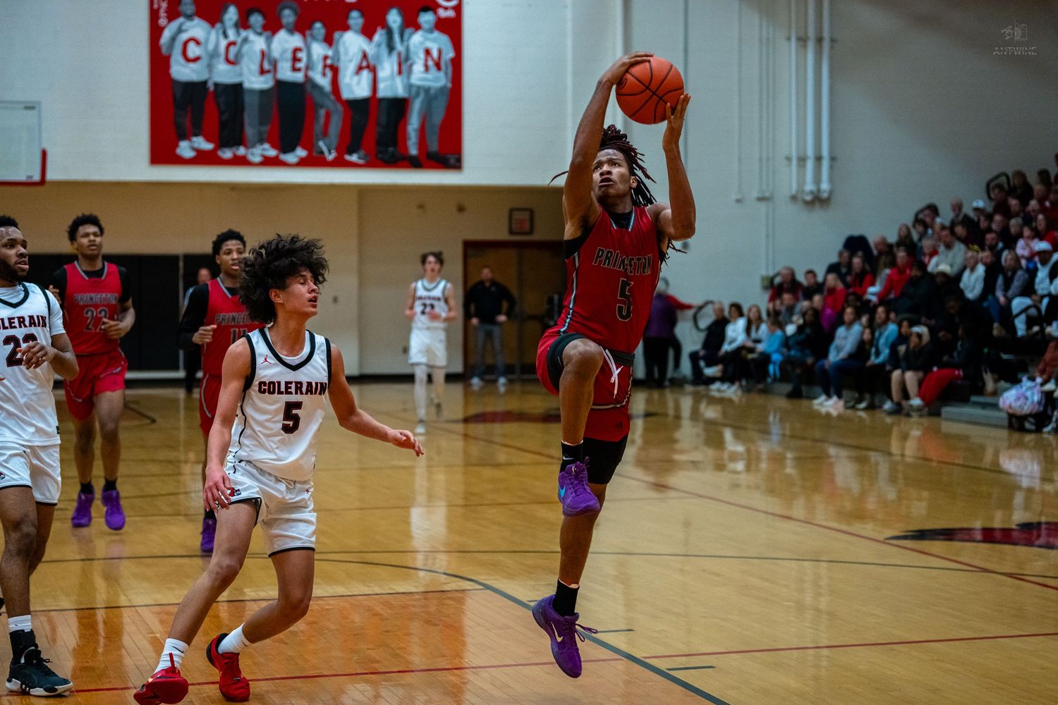 Basketball player jumps to catch a pass near the sideline during a high school gymnasium game.
